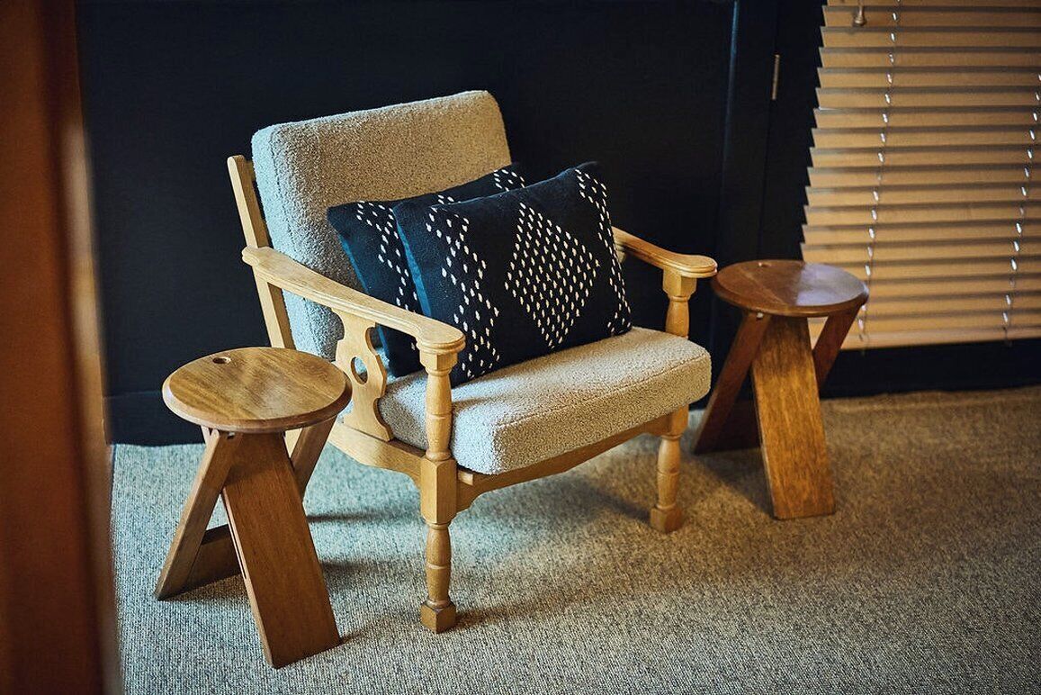 A wooden armchair with a cushion, two wooden side tables, and a window with blinds.