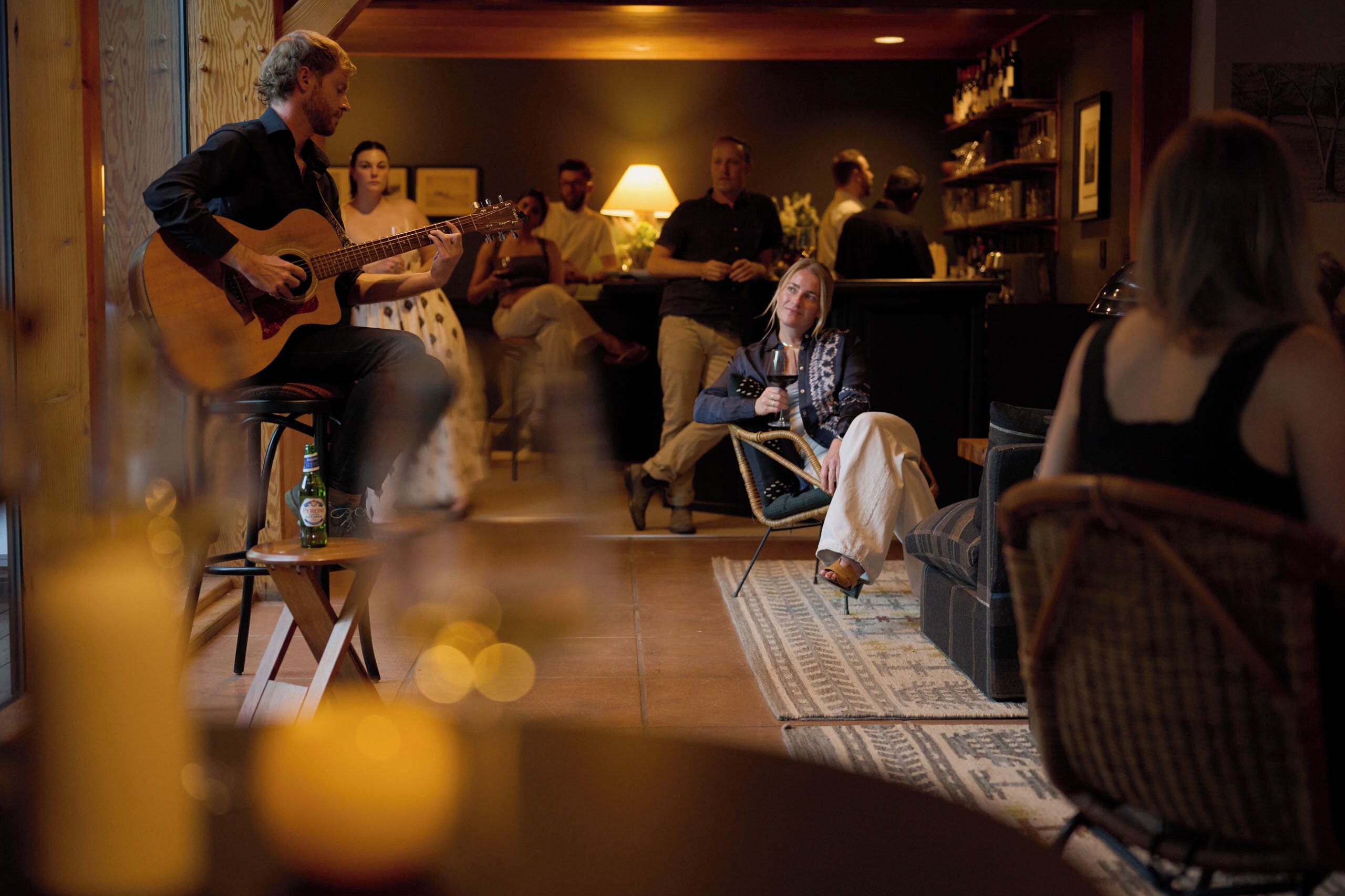 A musician performing in a cozy, dimly lit room with a small audience seated on sofas and chairs.