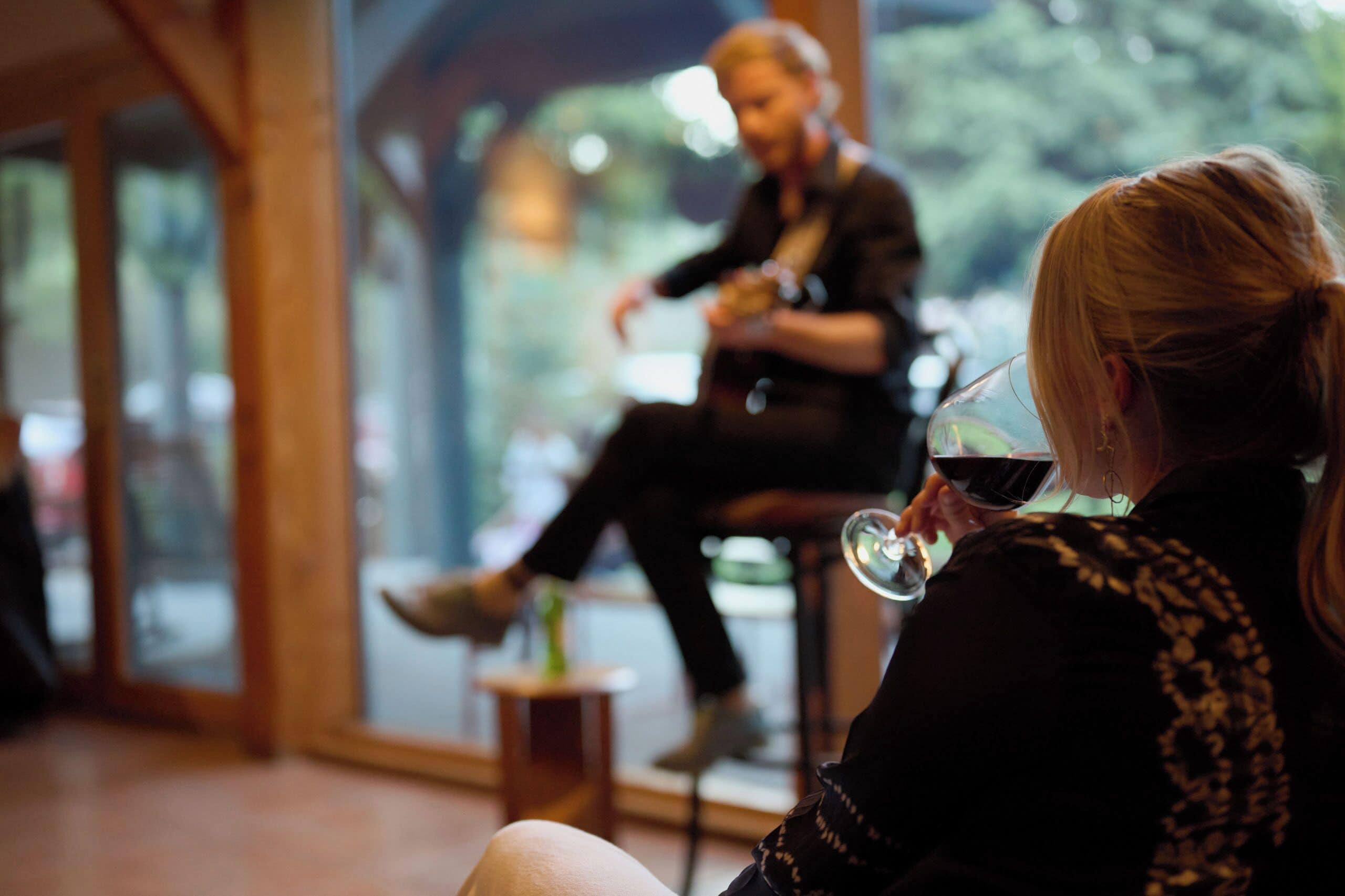 Une personne assise à l’intérieur, devant un verre de vin rouge, avec une fenêtre donnant sur un paysage verdoyant et un intérieur en bois.