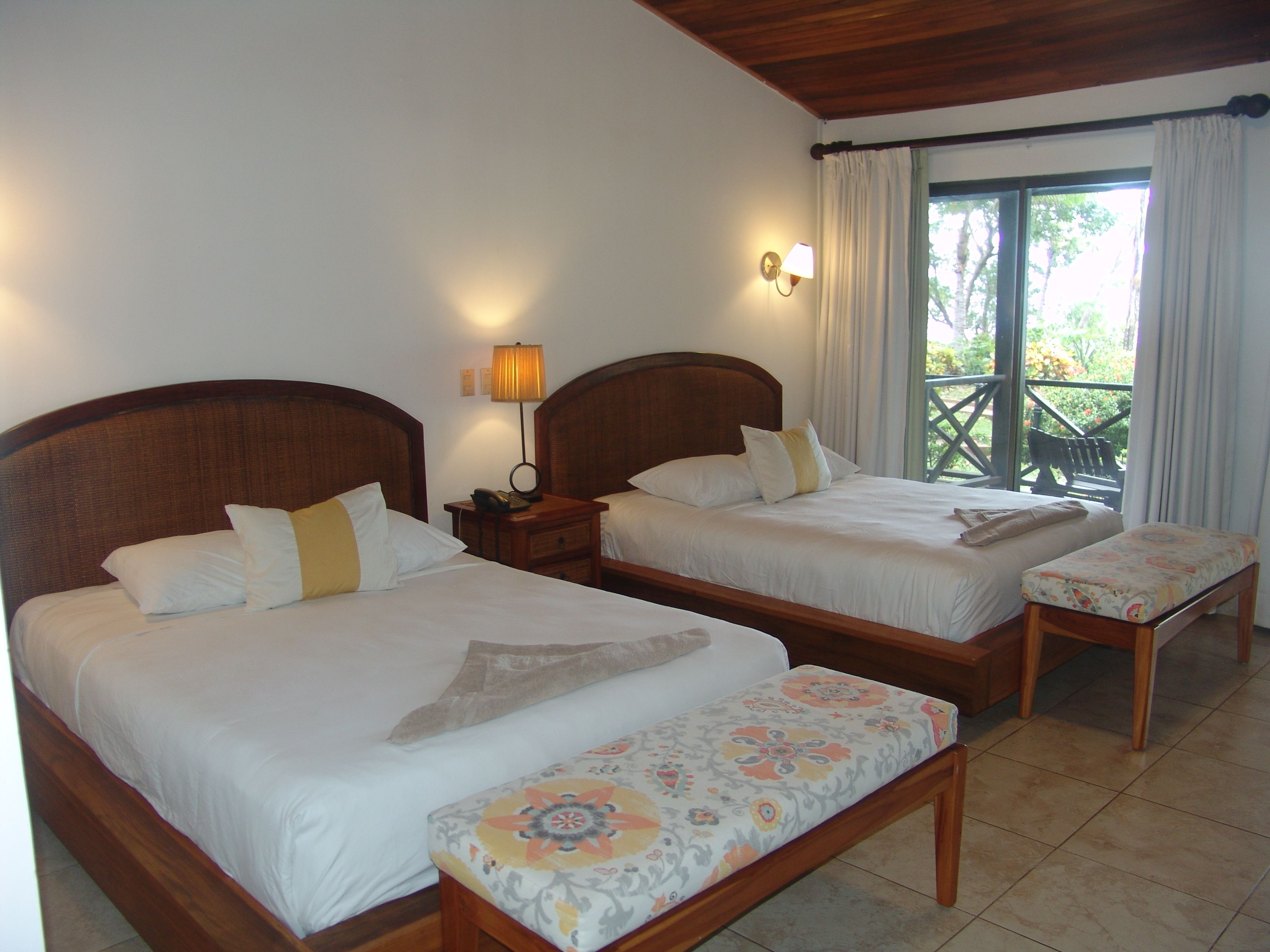 Habitación doble con dos camas, un banco, una mesita de noche con lámpara y vistas al exterior.