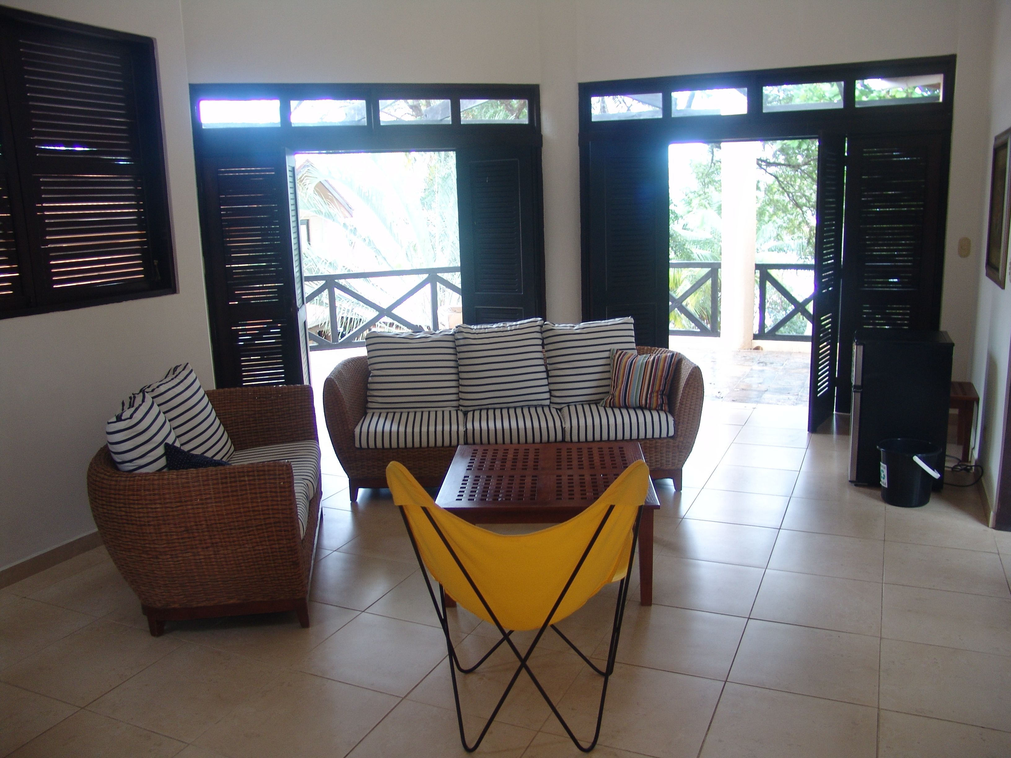 A living area with a sofa, chairs, and a table. There are windows with blinds, a speaker, and a tiled floor.