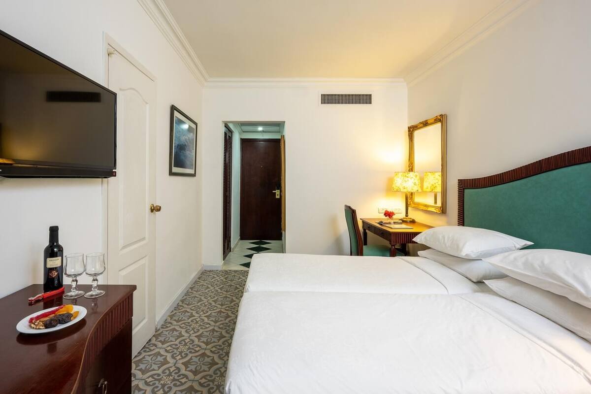 Habitación de hotel con una cama, un escritorio con lámpara, un televisor y una mesa de comedor con una botella y vasos.