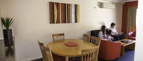 Una habitación con una mesa redonda de madera, cuatro sillas, un sofá y un sillón rojo. Hay un cuadro en la pared y un equipo de aire acondicionado sobre el sofá.