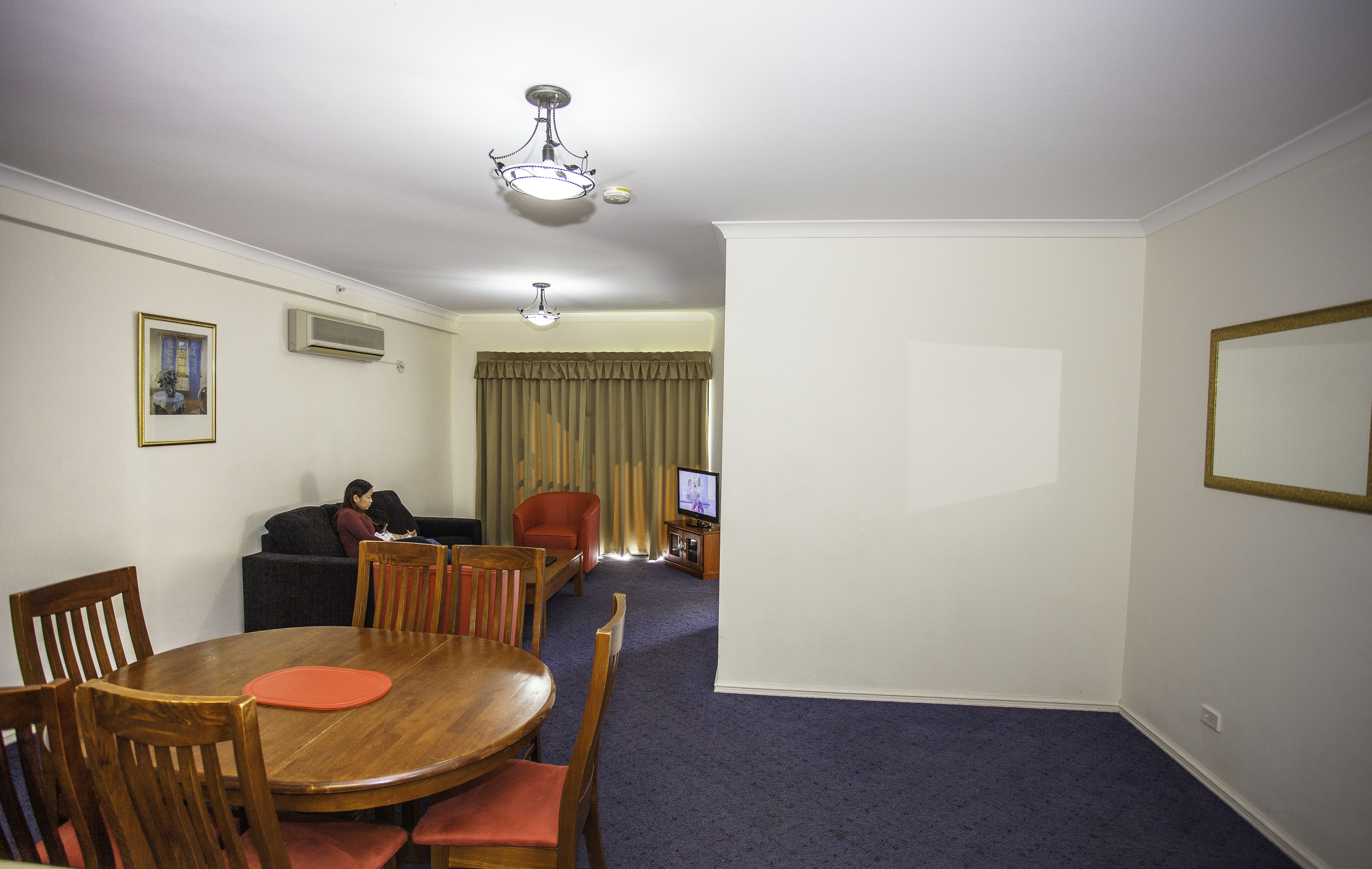 A hotel room with a dining table, chairs, a sofa, a television, and a ceiling light fixture.