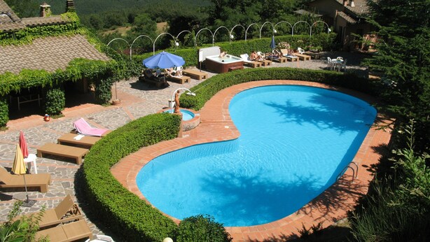 Una piscina de borde curvo, rodeada de sillas de descanso y sombrillas, con colinas y un cielo despejado al fondo.