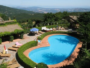 Piscina com borda curva, rodeada por espreguiçadeiras e guarda-sóis, com colinas e céu claro ao fundo.