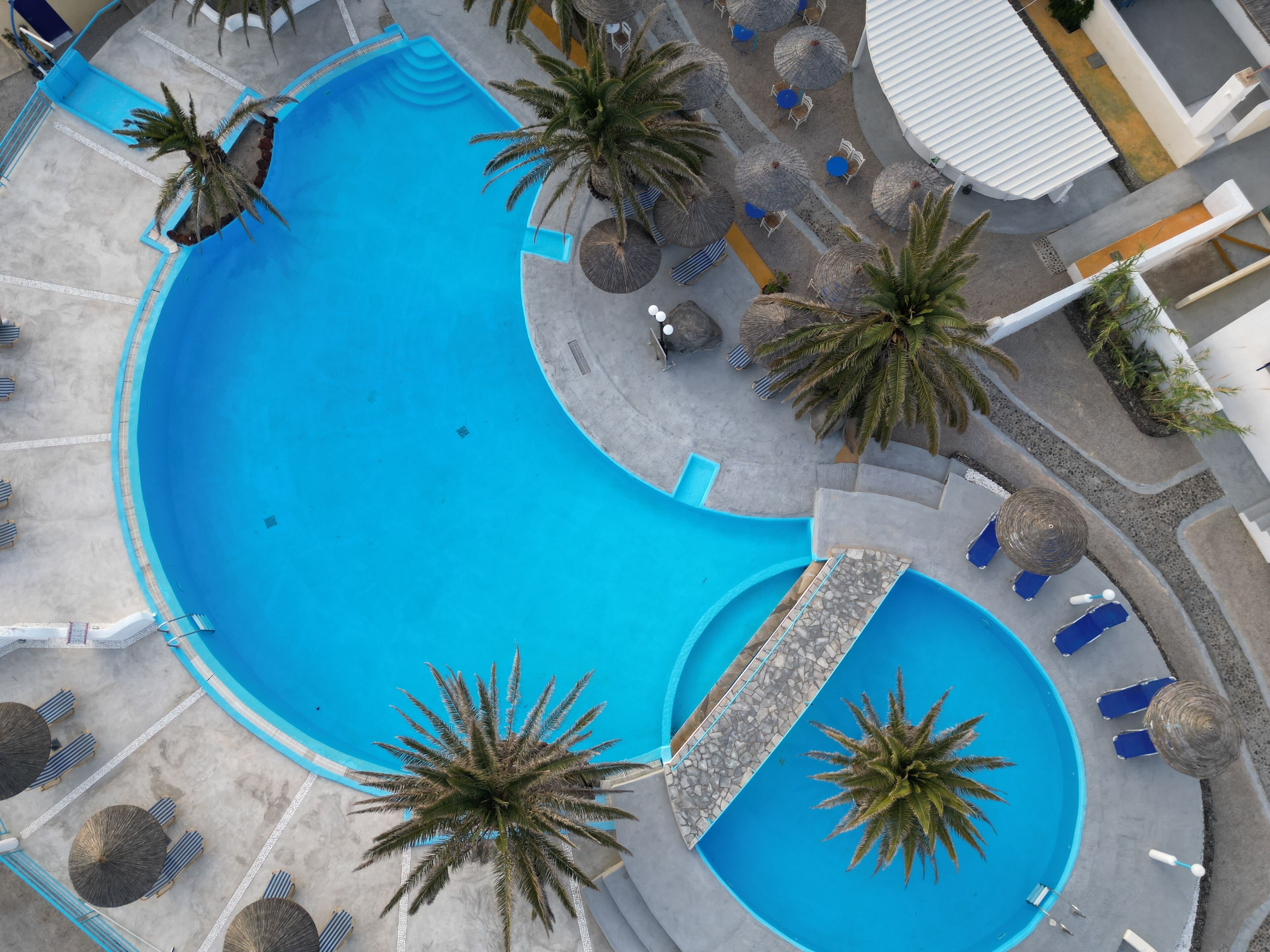 Vue aérienne d’une piscine à l’eau bleue, entourée de palmiers et de chaises longues.