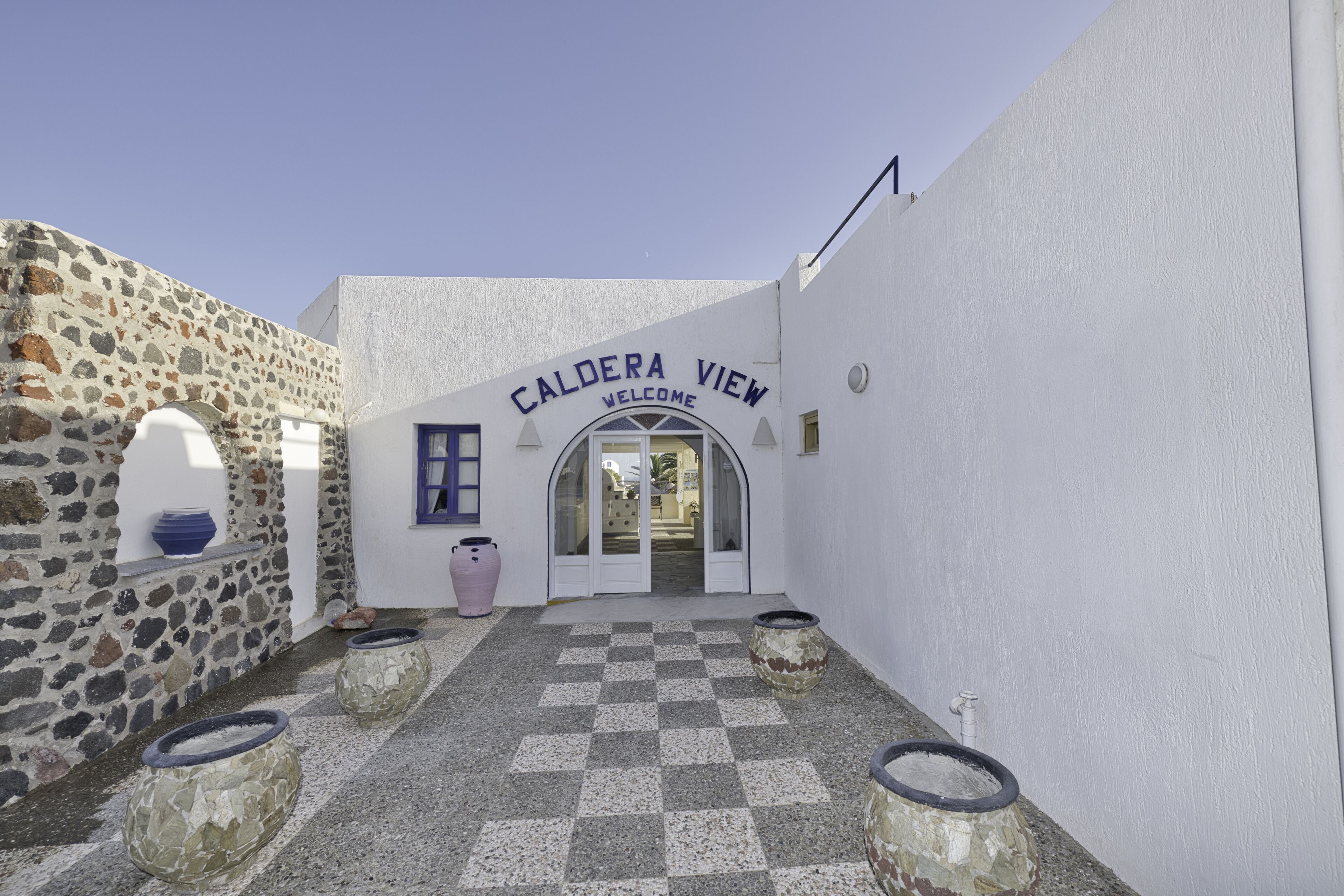 A courtyard with a stone wall, a checkered floor, and a sign reading "Caldera View Welcome".