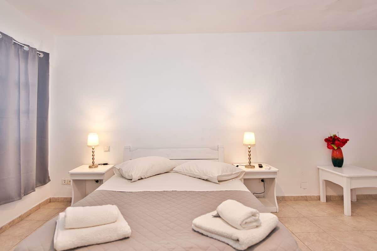 A hotel room with a bed, two bedside lamps, a nightstand with a vase of red flowers, and a window with grey curtains.