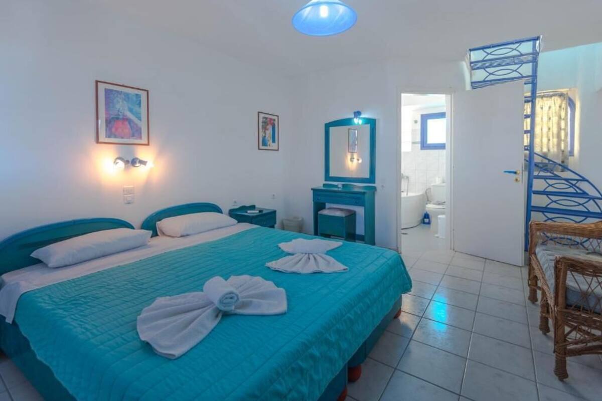 A hotel room with a bed, a nightstand, a mirror, a bathroom visible through an open door, and a staircase with a blue railing.