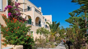 A building with multiple balconies, arched windows, and a pathway lined with trees and shrubs. - Aelia by Eltheon (Santorini)