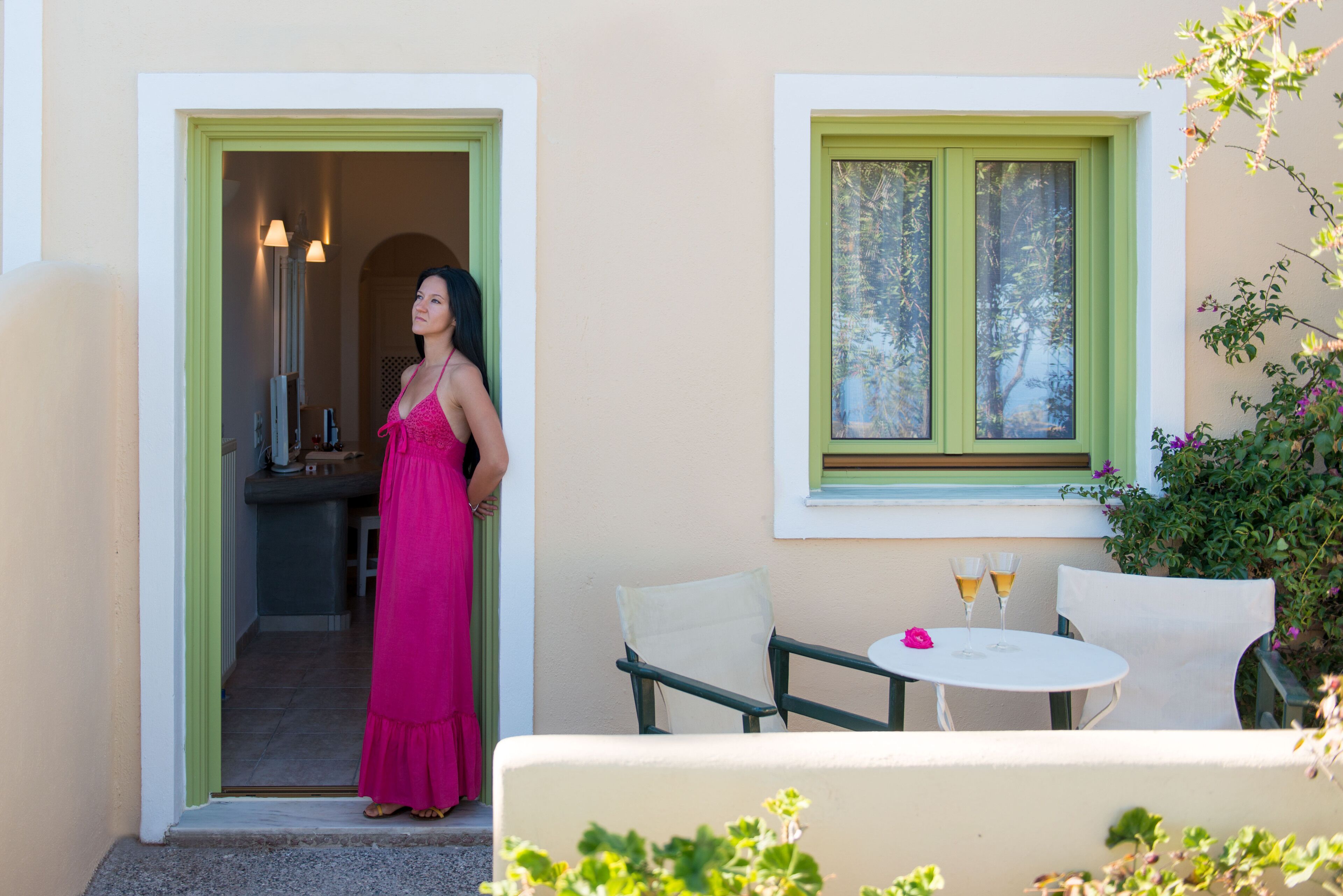 A woman in a pink dress standing at the entrance of a building with a green door and a window with a view of trees.