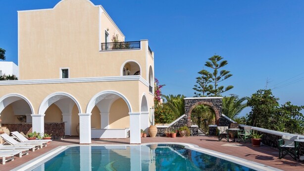 A swimming pool with a clear blue water surface, surrounded by white columns and a beige building with balconies. - Aelia by Eltheon (Santorini)