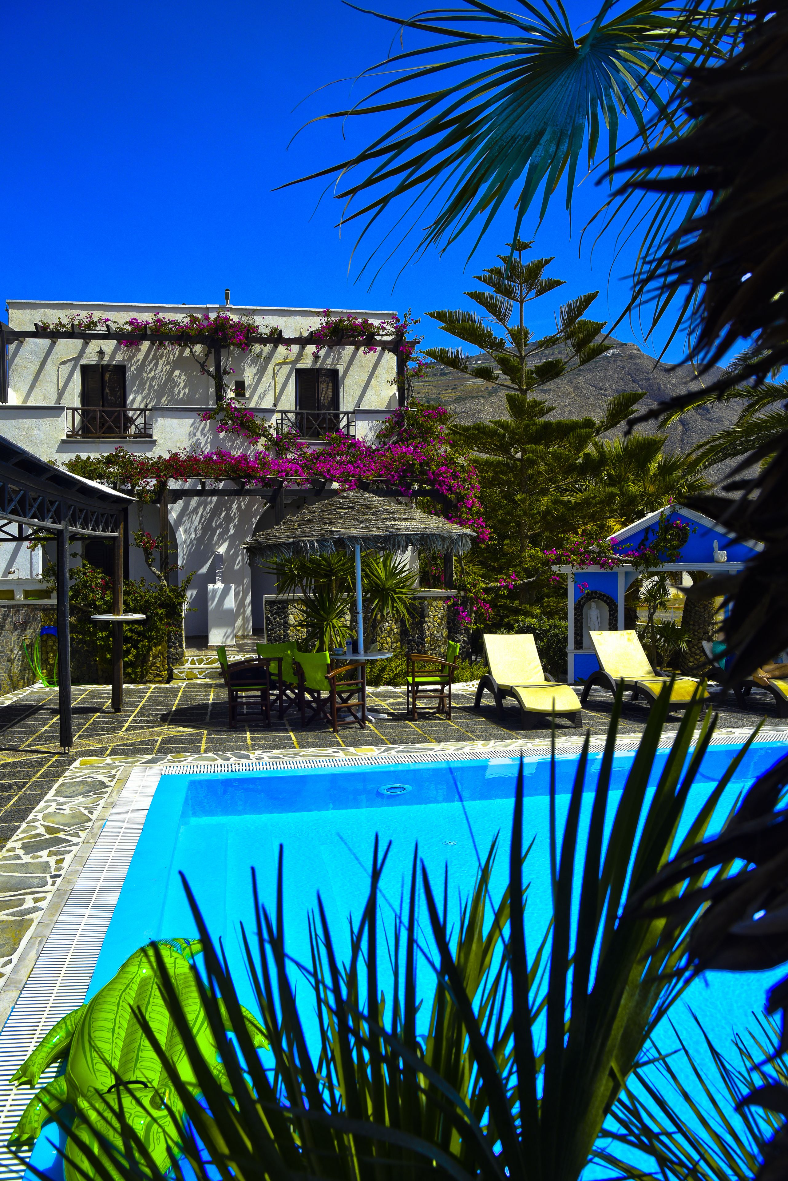 Un área de piscina con sillones reclinables, una pérgola con flores y un edificio de fachada blanca y ventanas.