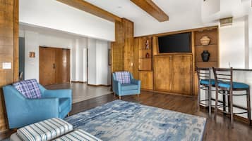 Amplia sala de estar con un sillón azul, un reposapiés a rayas y una mesa de centro de madera. Se observa un mueble de madera con estantes y un televisor de pantalla plana montado encima.