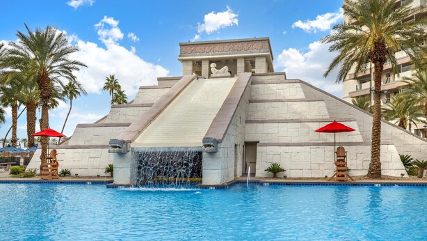 A large swimming pool with a waterfall feature, palm trees, and a building with hieroglyphic carvings.