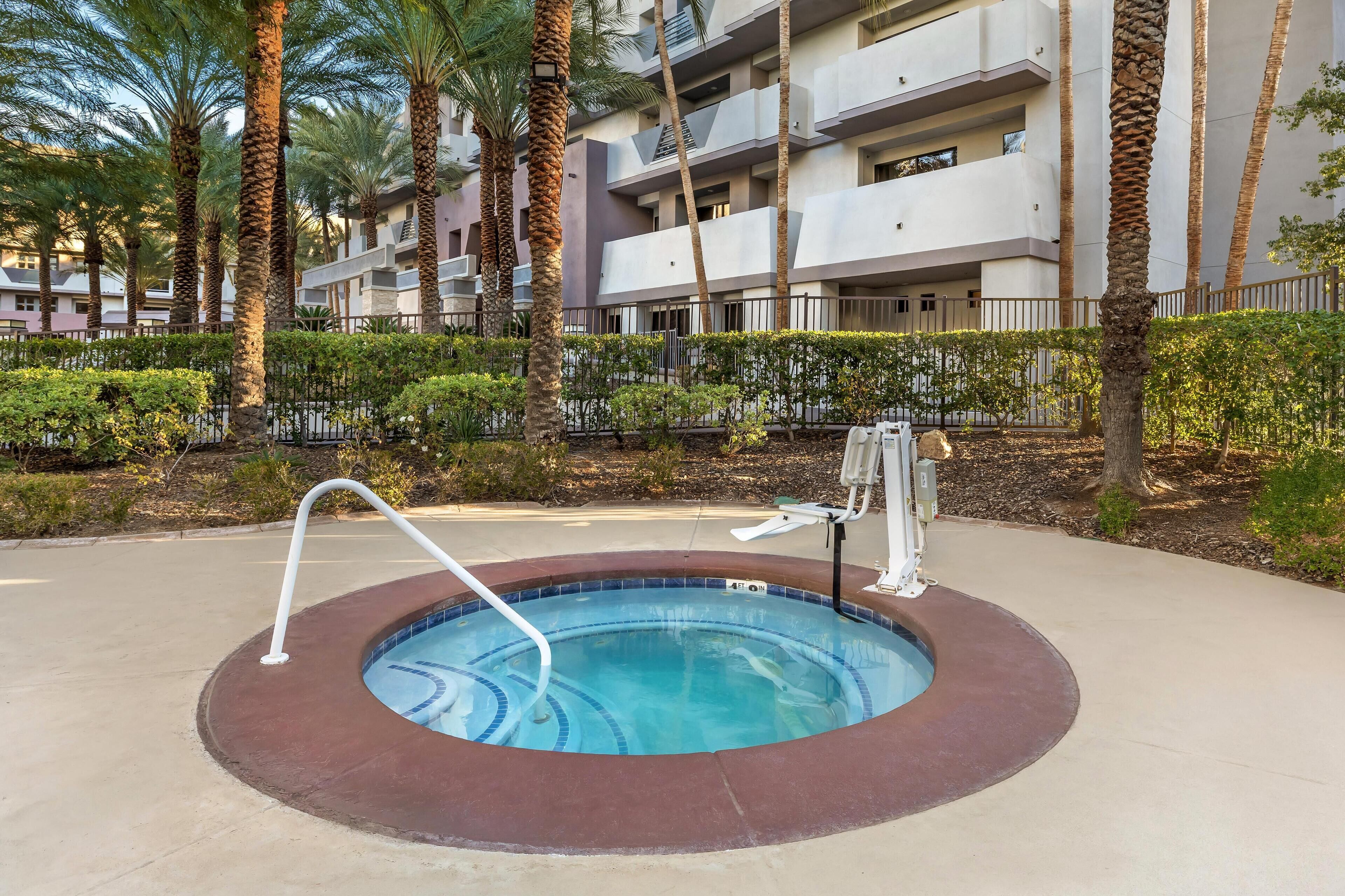 Une piscine avec un jacuzzi, entourée de palmiers et d’une clôture, avec un immeuble à plusieurs étages en arrière-plan.