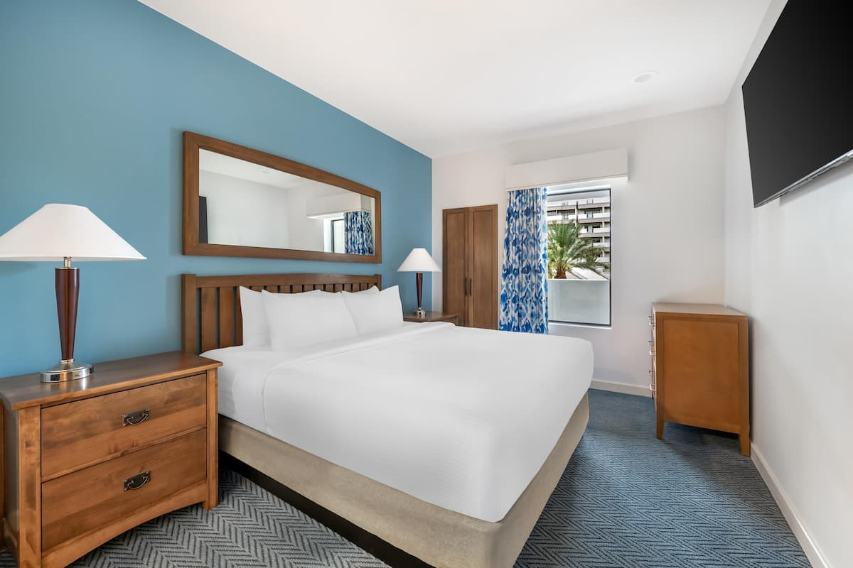 Une chambre d’hôtel avec un grand lit, une table de chevet en bois, deux lampes, un miroir, une armoire et un téléviseur.