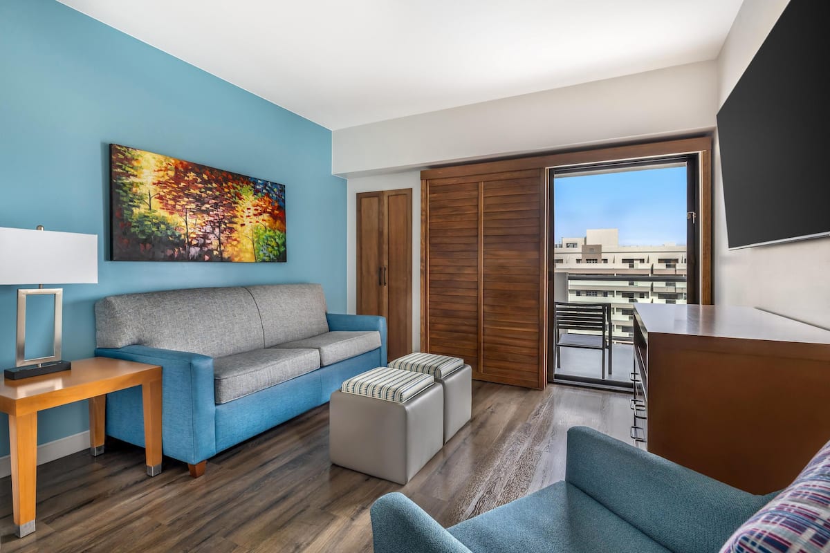 Une chambre d’hôtel moderne avec un mur bleu, un canapé gris, une table en bois et une vue sur la ville grâce à de grandes fenêtres.