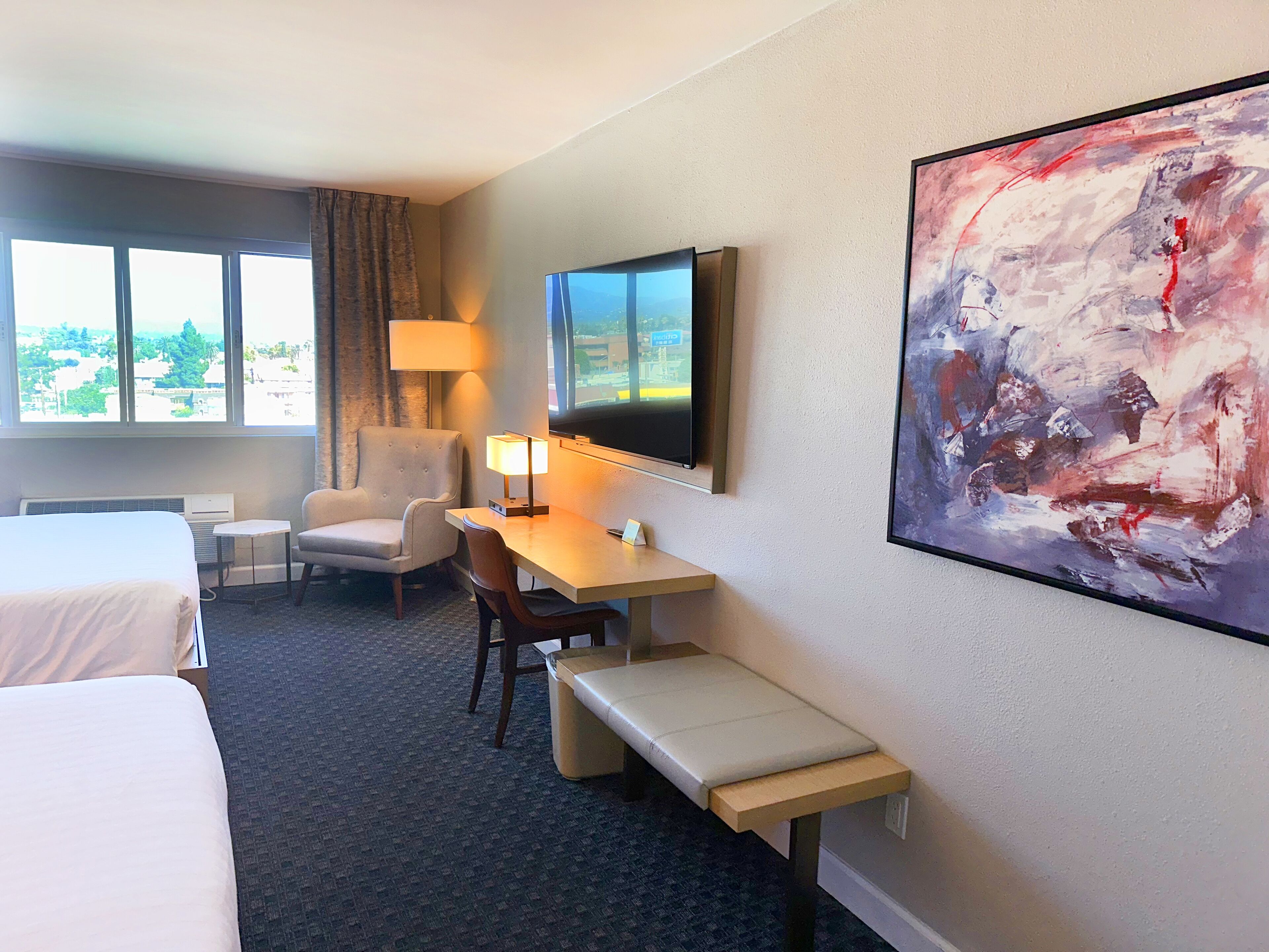 A hotel room with a bed, a desk with a chair, a television, and a large abstract painting on the wall.