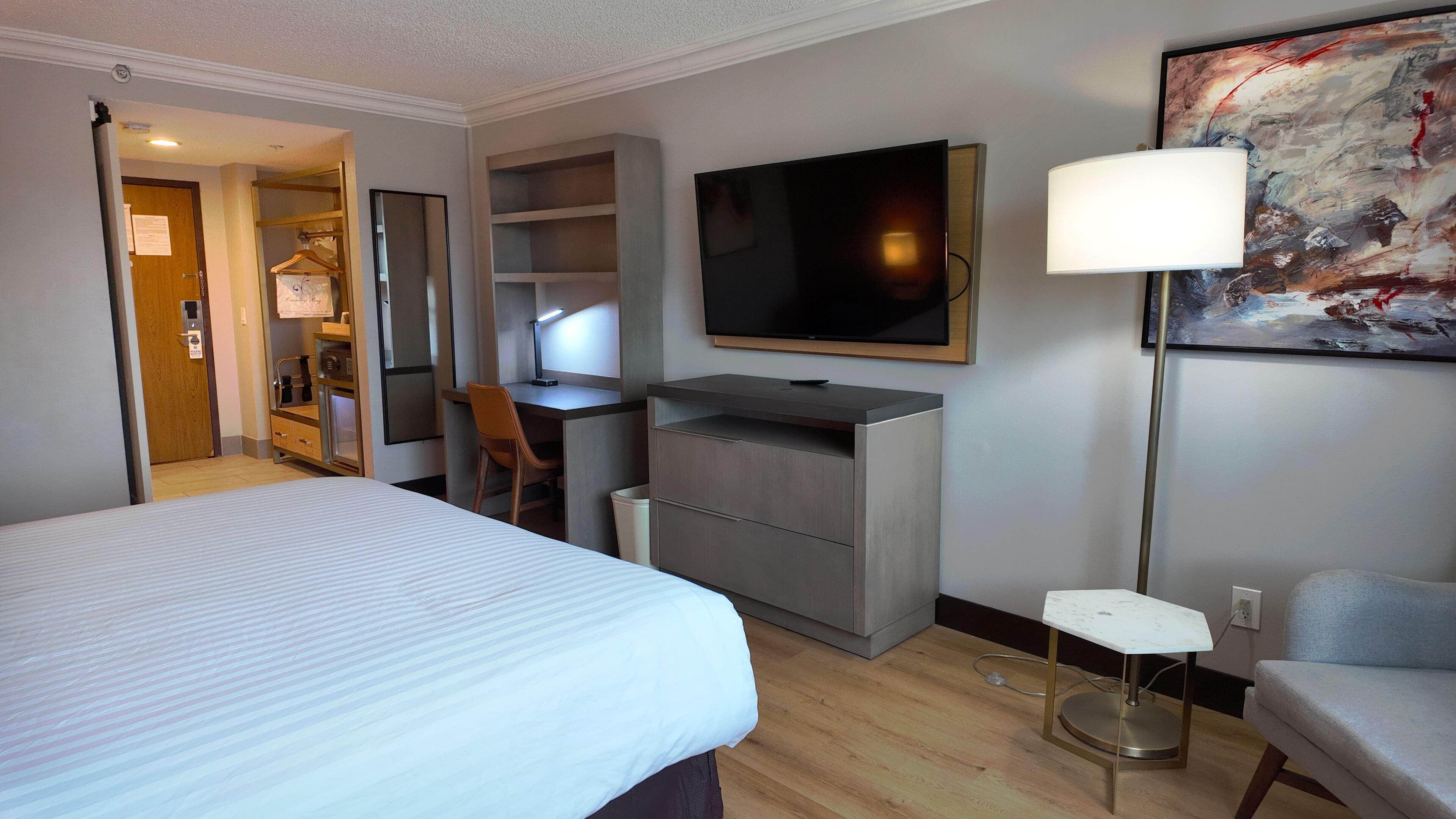 A hotel room with a bed, desk, chair, TV, and a painting on the wall.