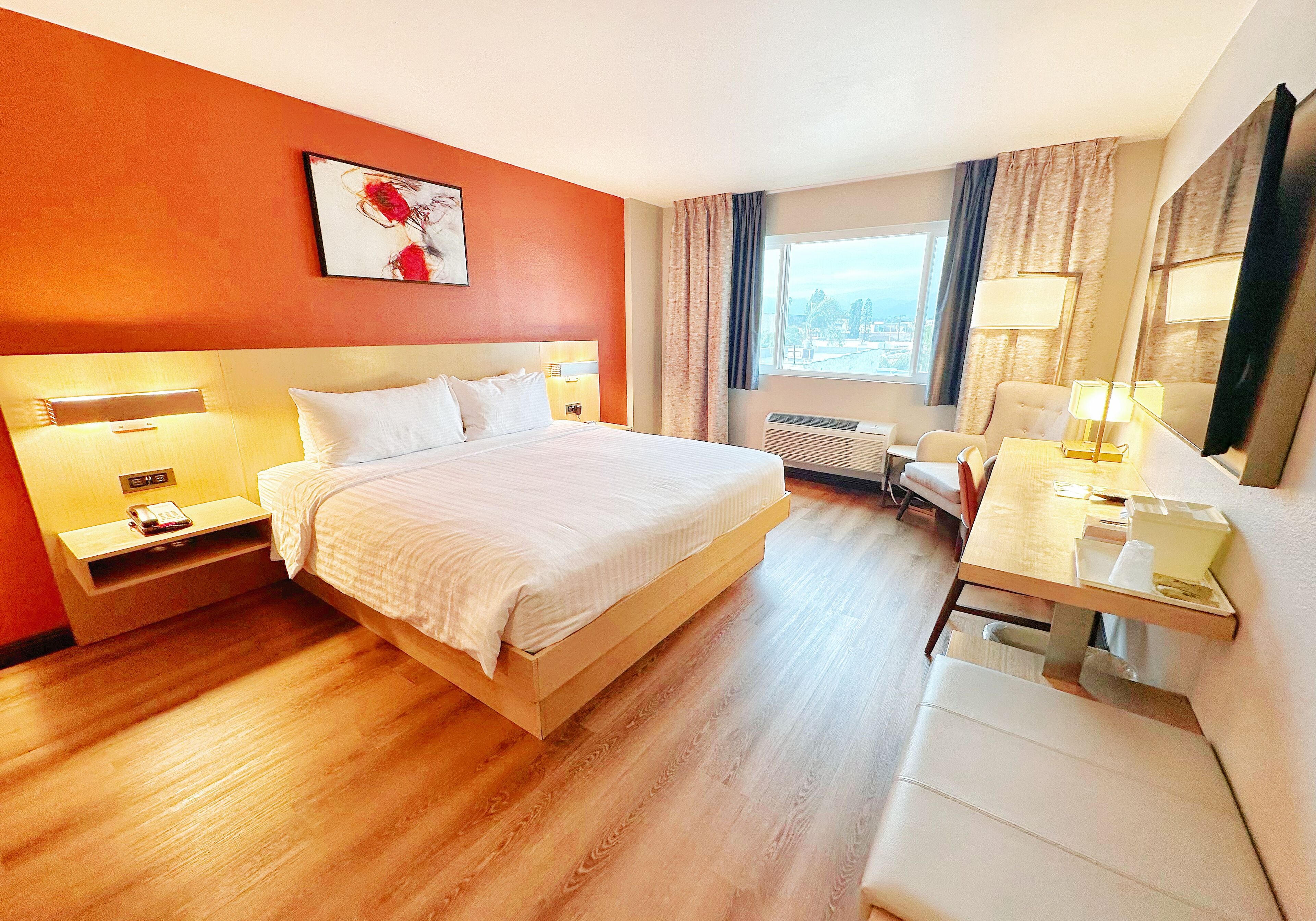 A hotel room with a bed, bedside tables, a desk, a chair, a window with curtains, and a city view.