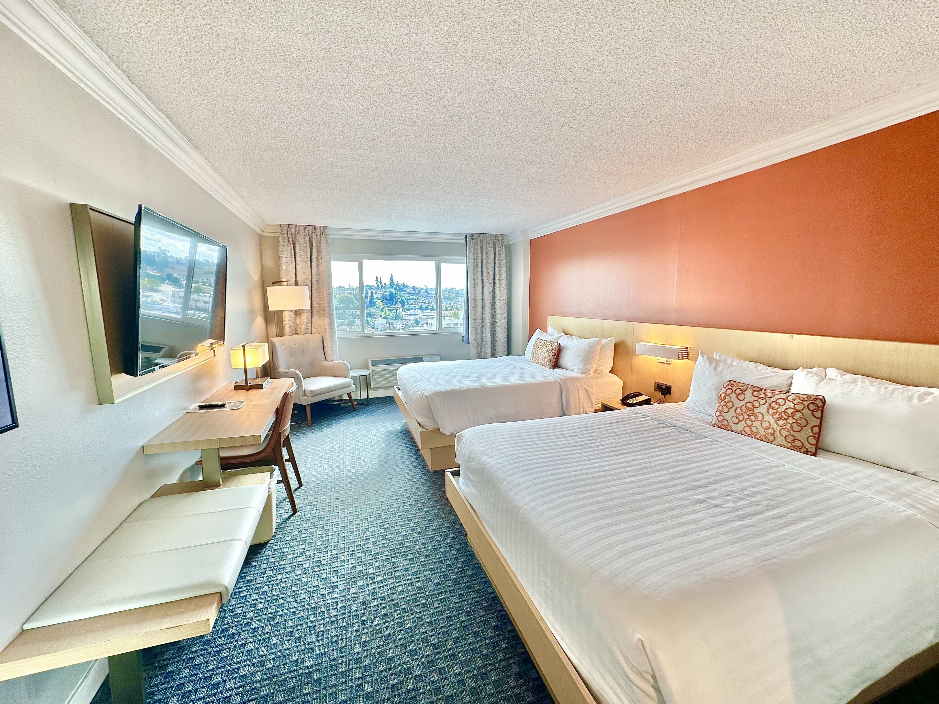 A hotel room with two beds, a desk, a chair, a TV, and a window with a city view.