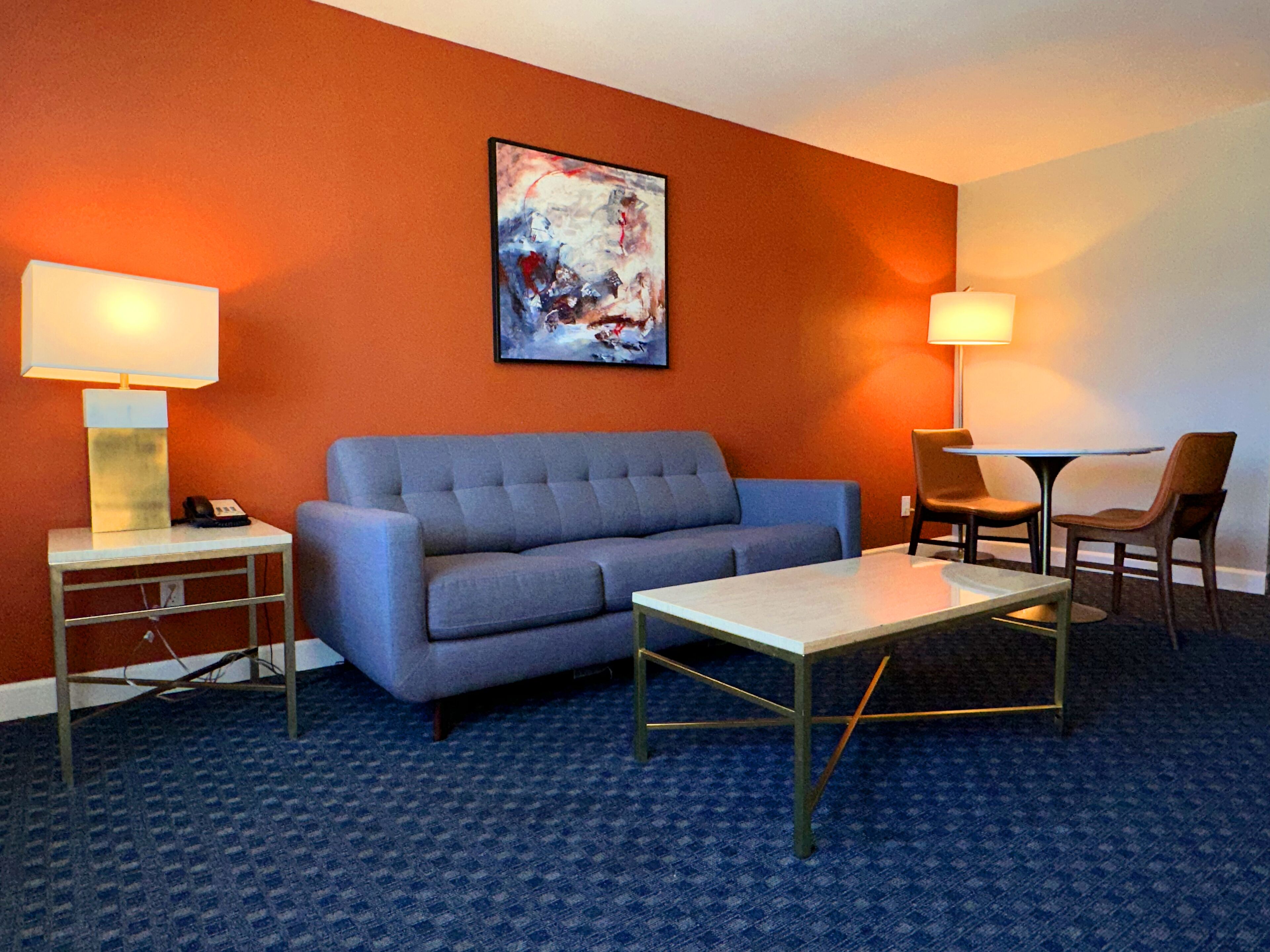 A hotel room with a blue sofa, a small table, two chairs, a lamp, and a painting on the wall.