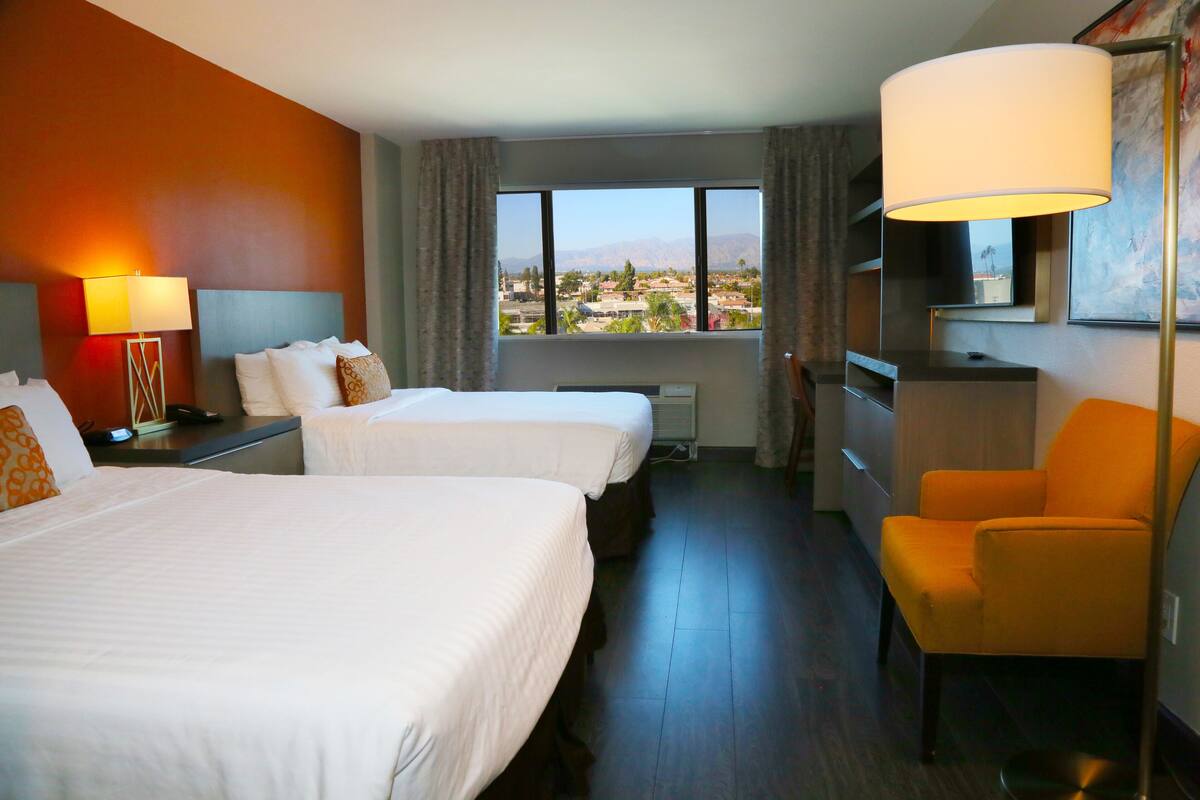 A hotel room with two beds, a desk, a chair, a TV, and a large window with a view of a city and mountains.