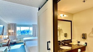 Une chambre d’hôtel comprenant un lit, un bureau, une chaise, une salle de bain avec un lavabo, un miroir et des articles de toilette.