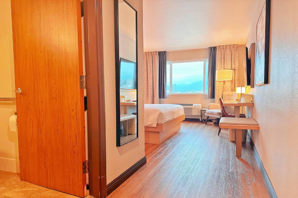 A hotel room with a wooden door, a bed, a desk with a chair, a window with curtains, and a lamp.