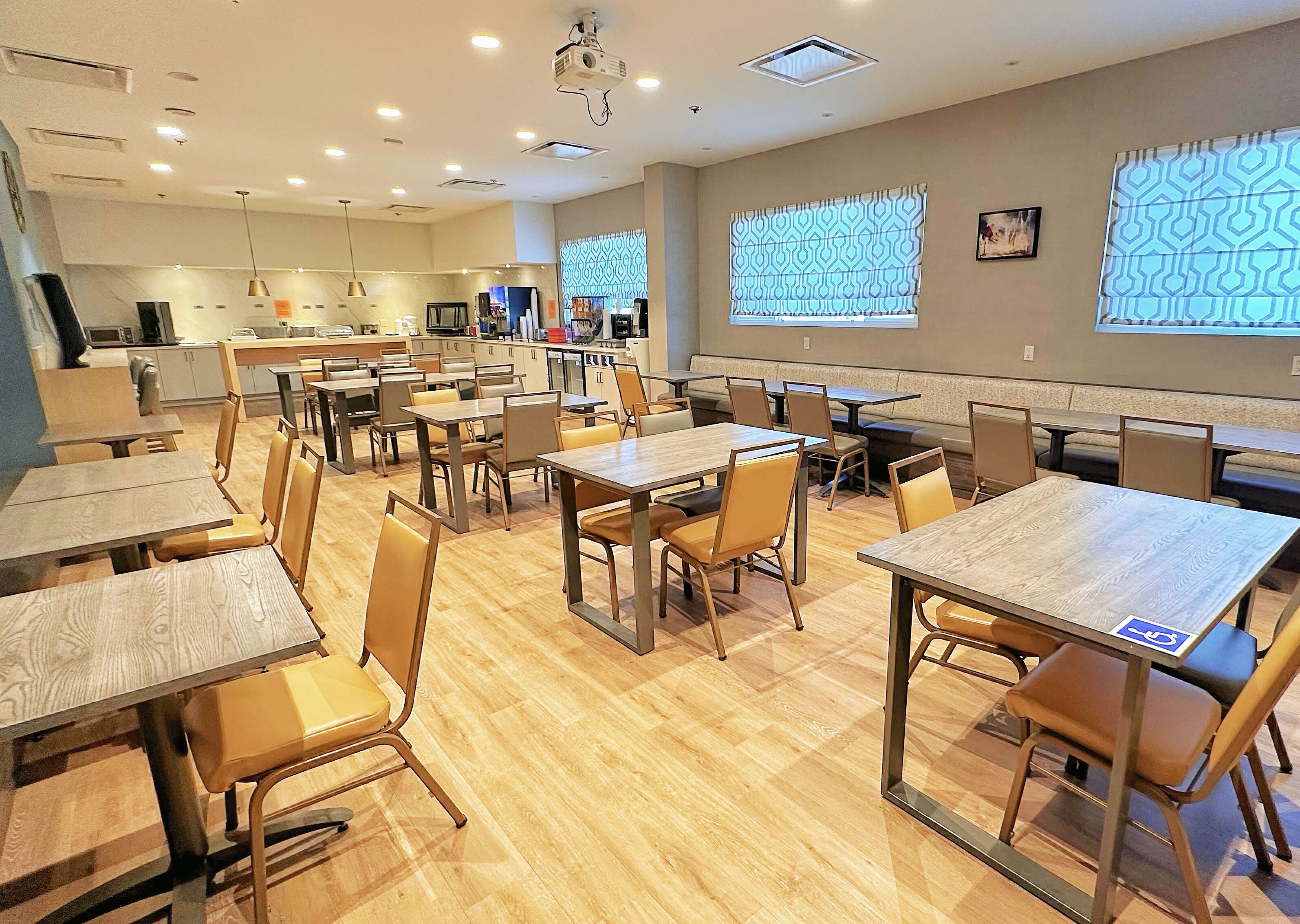 A spacious dining area with multiple tables and chairs, a kitchen in the background, and patterned windows.