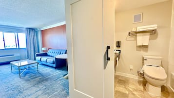 A hotel room with a living area featuring a sofa and a coffee table, a bathroom with a toilet and a shower, and a door leading to another room.
