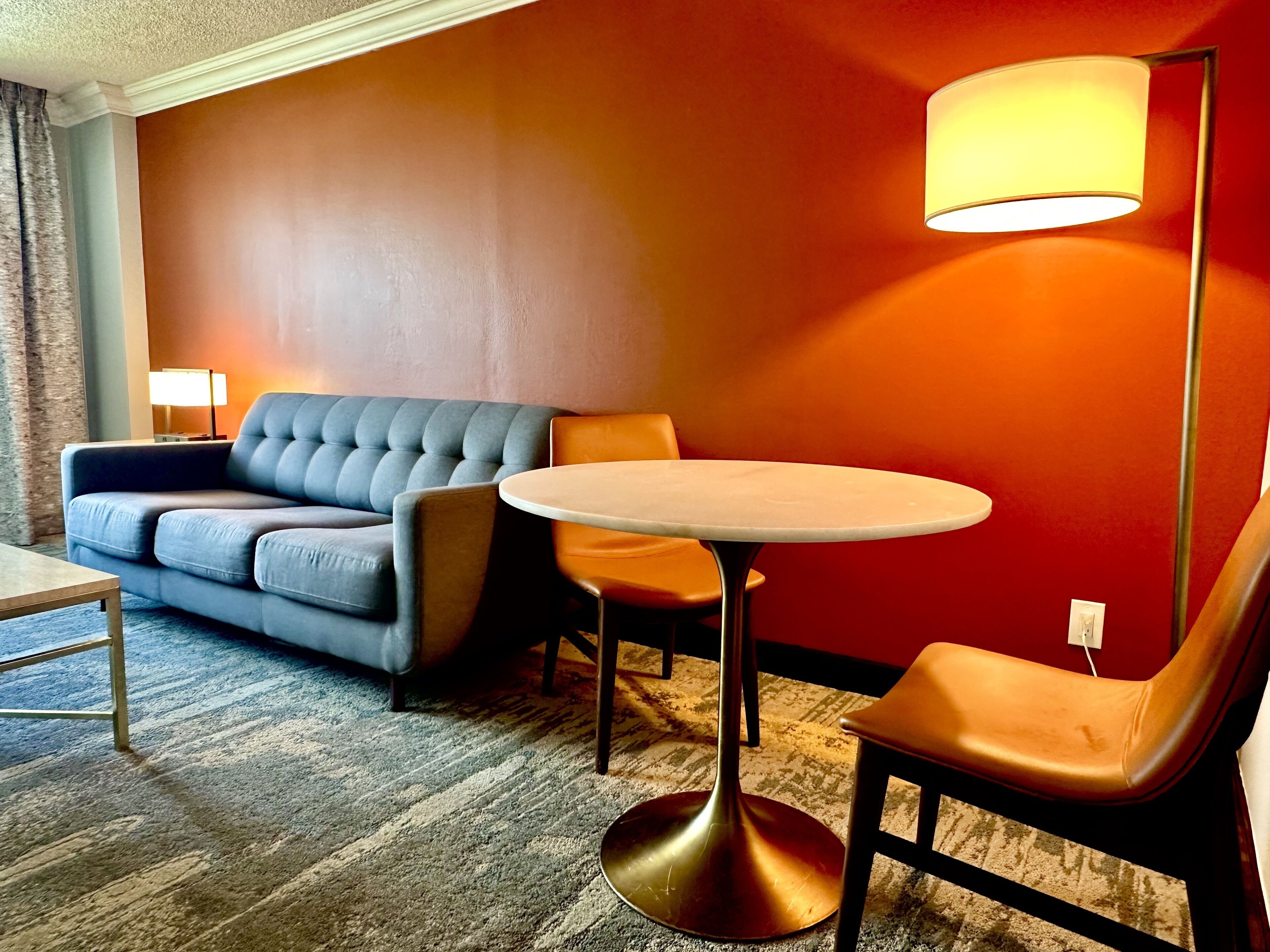 A modern hotel room with a grey sofa, a round table, and a single orange chair.