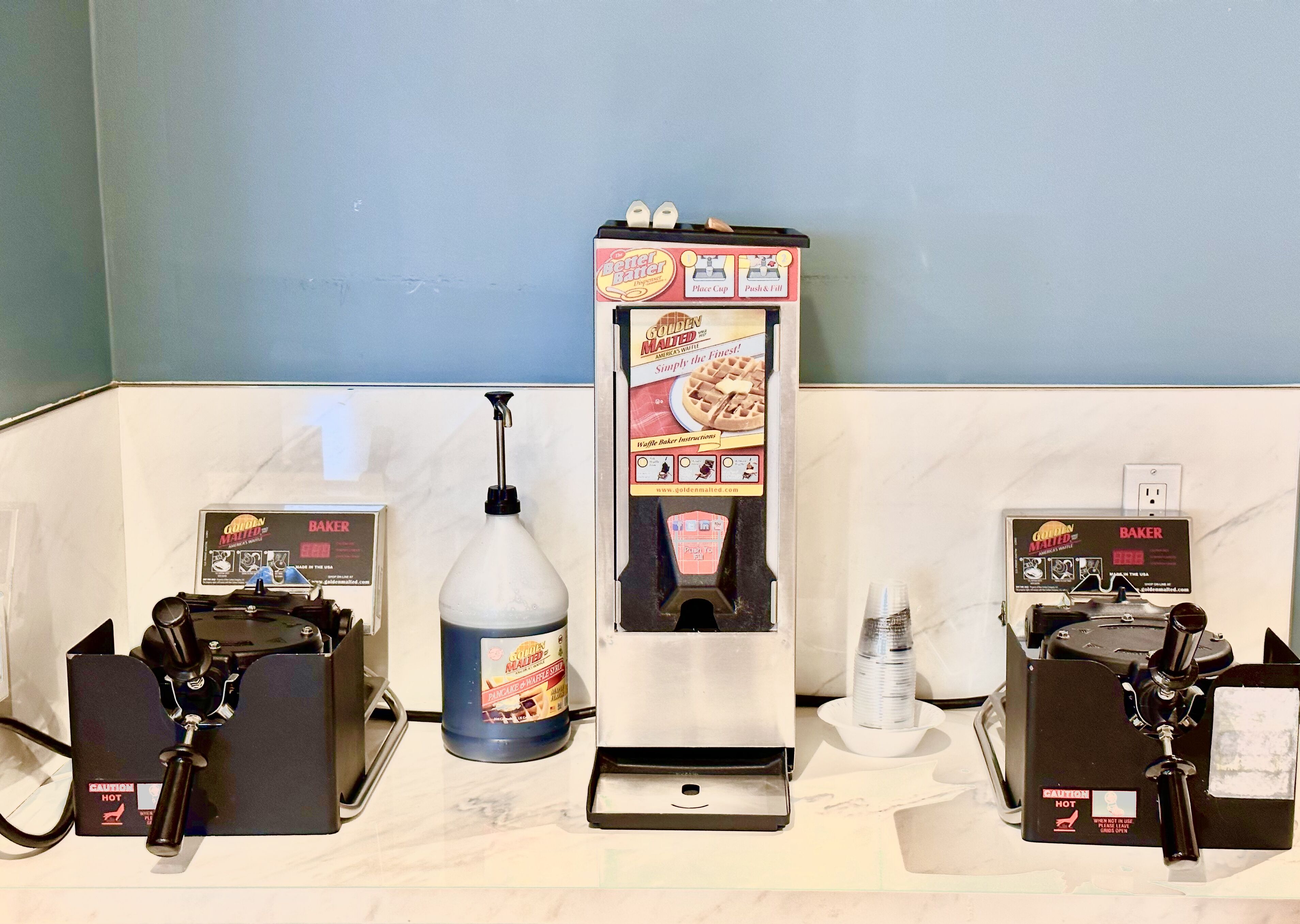 A breakfast bar with waffle makers, a bottle of syrup, and a waffle maker box.