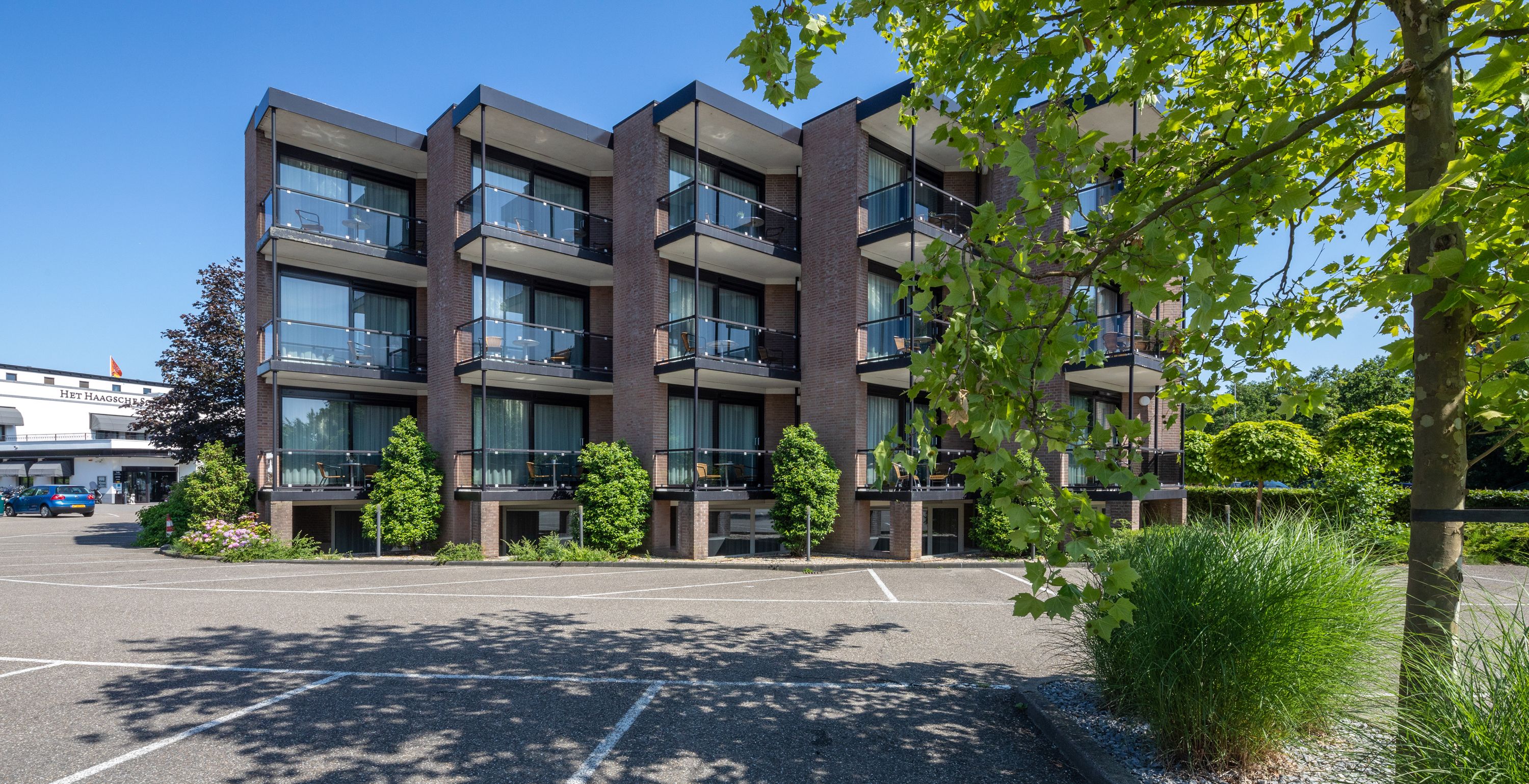 Een meerlaags gebouw met balkons, een parkeerplaats en omliggend groen.