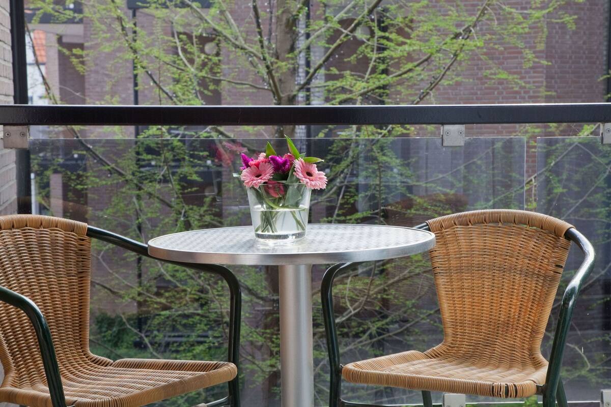 Une table ronde, deux chaises en osier et un vase de fleurs sont disposés sur un balcon doté d’une balustrade en verre.