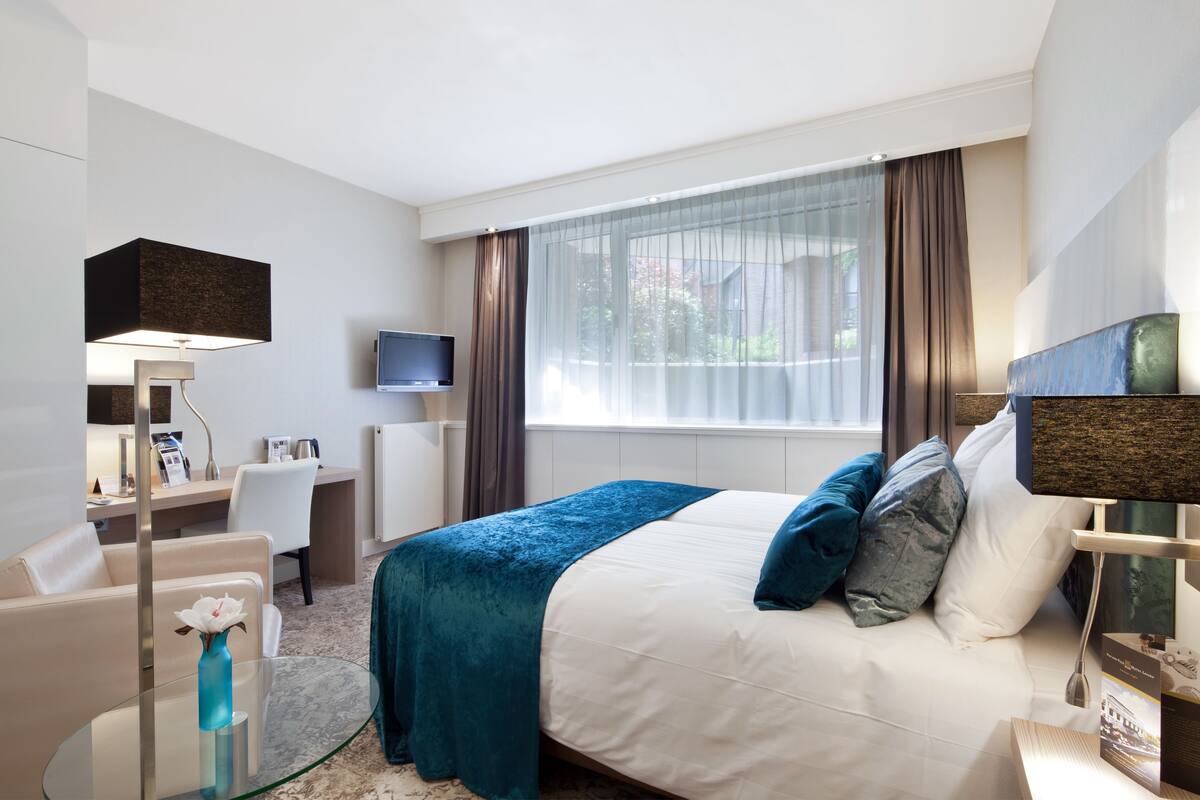 Une chambre d’hôtel avec un lit, un bureau, une chaise, une télévision et une fenêtre avec des rideaux.