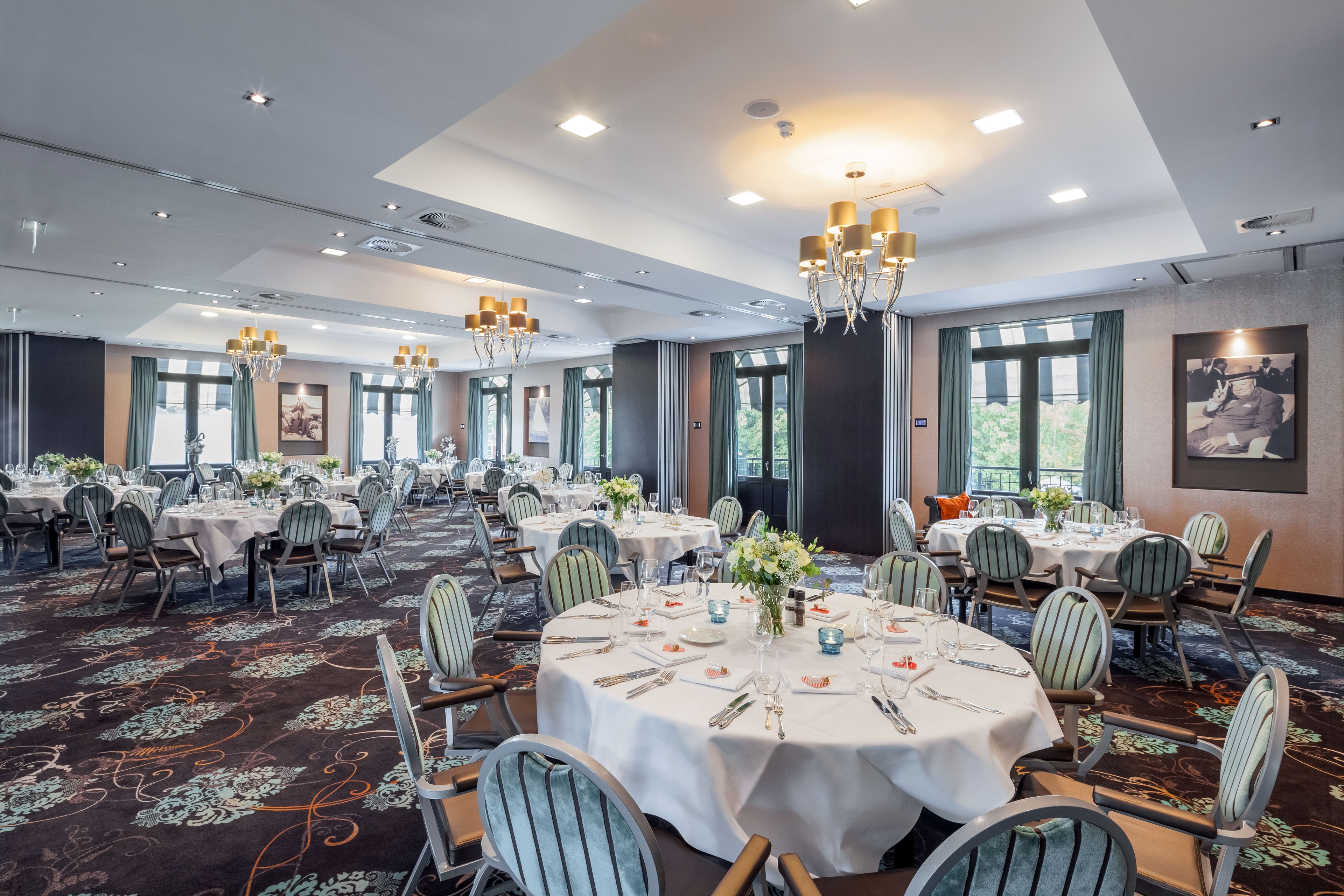 Een ruime balzaal met ronde tafels, stoelen, kroonluchters en grote ramen.