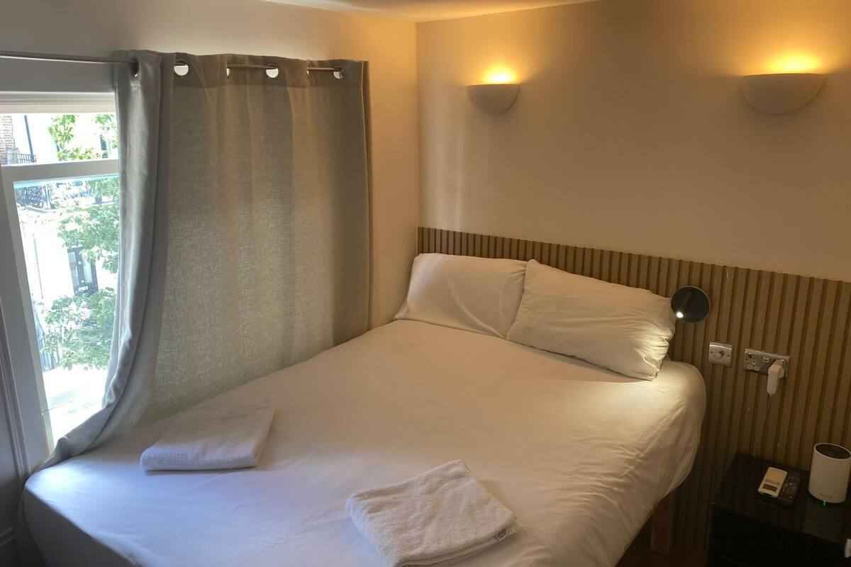 Een opgemaakt bed met witte lakens, een raam met gordijnen en een nachtkastje met een lamp en een telefoon.