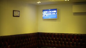 A corner booth with a cushioned backrest, a glass table with a flower, a framed picture on the wall, and a digital screen displaying a map. - Exhibition Court Hotel 4 (London)