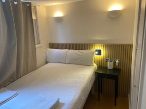 A hotel room with a bed, two wall-mounted lights, a bedside table with a phone and a cup, and a window with curtains. - Exhibition Court Hotel 4 (London)