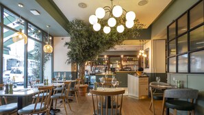 A restaurant interior with wooden tables and chairs, a central plant, and a bar area.