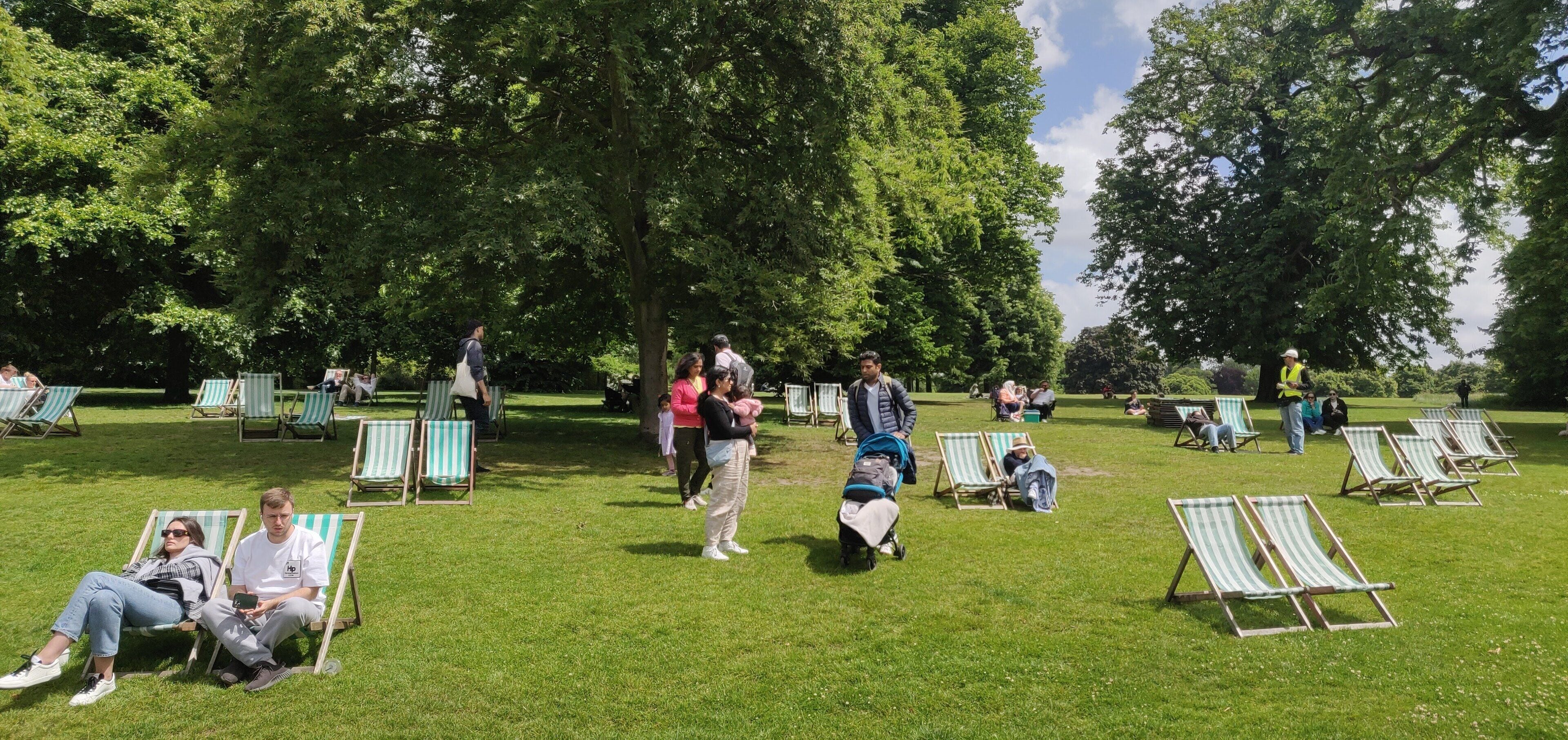 A park with green grass and trees, people sitting on lawn chairs, and a person in a yellow vest.