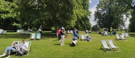 A park with green grass and trees, people sitting on lawn chairs, and a person in a yellow vest.
