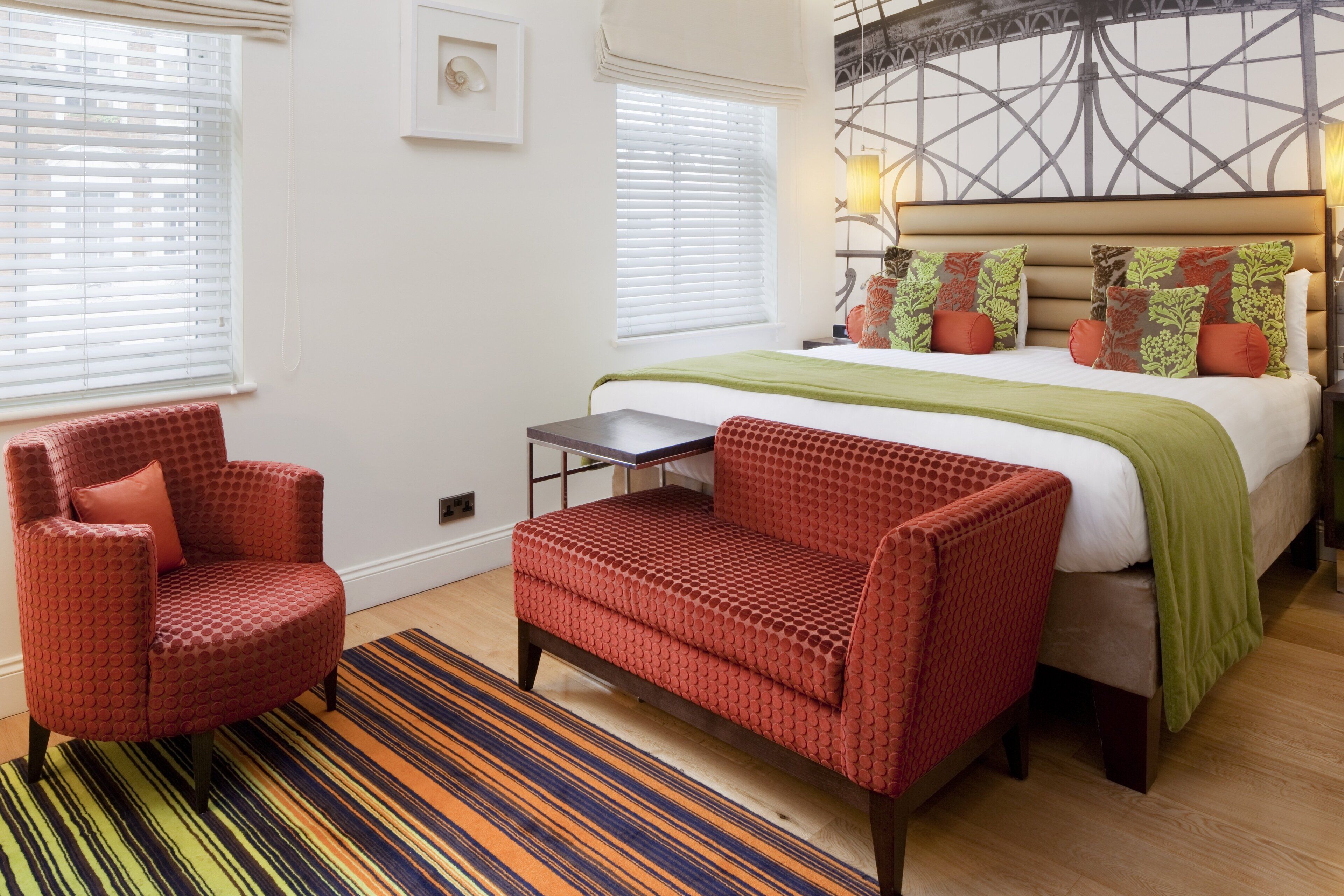A hotel room with a bed, two red armchairs, a small table, and a colorful striped rug.