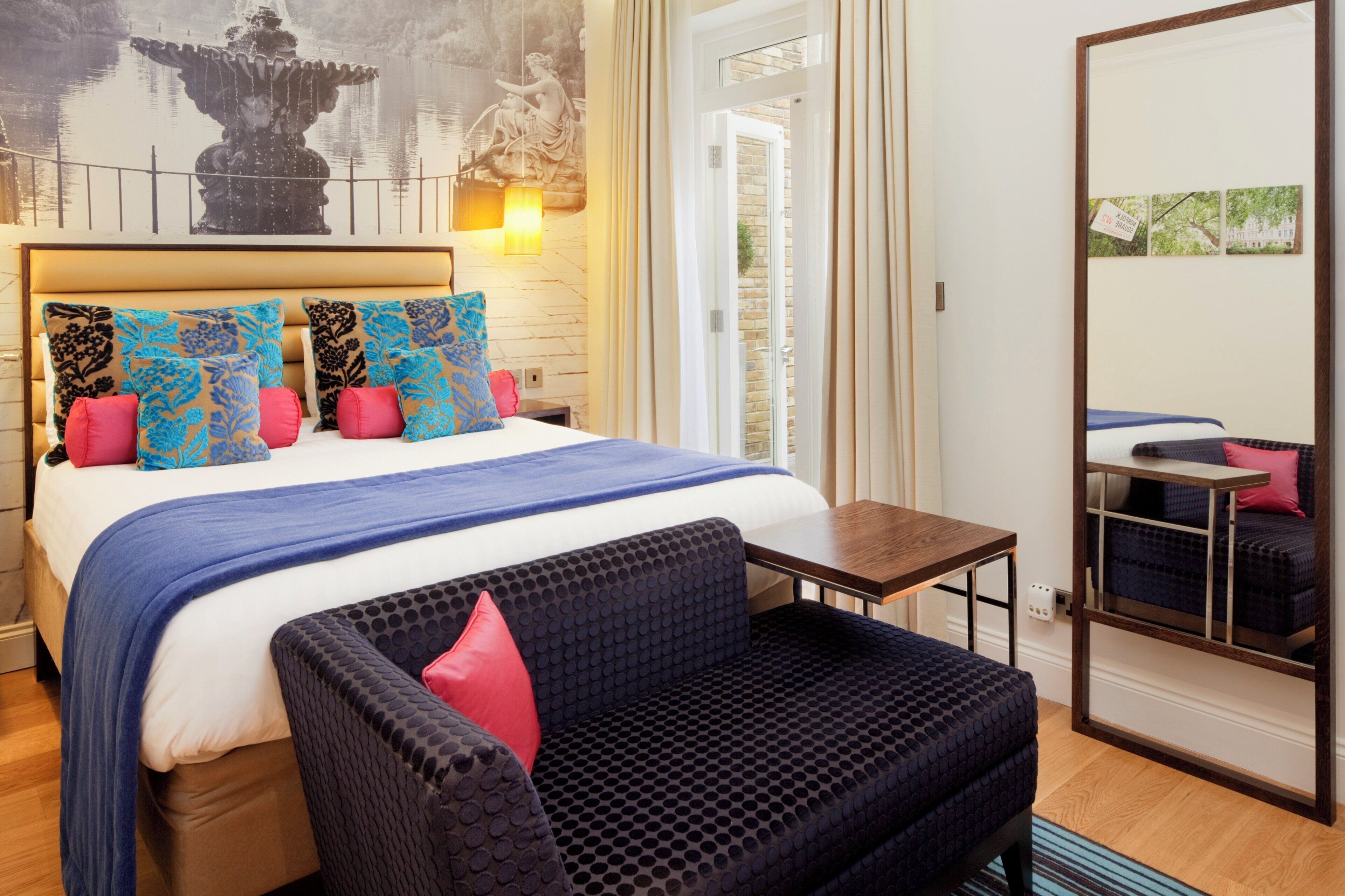 A hotel room with a bed, a sofa, a small table, a mirror, and a wall with a fountain and a statue.