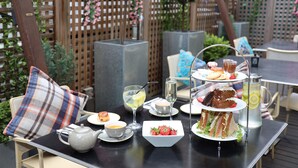A table set with a variety of food and drinks, including sandwiches, pastries, and a pitcher of lemonade, on an outdoor patio with wooden lattice fencing and greenery in the background.