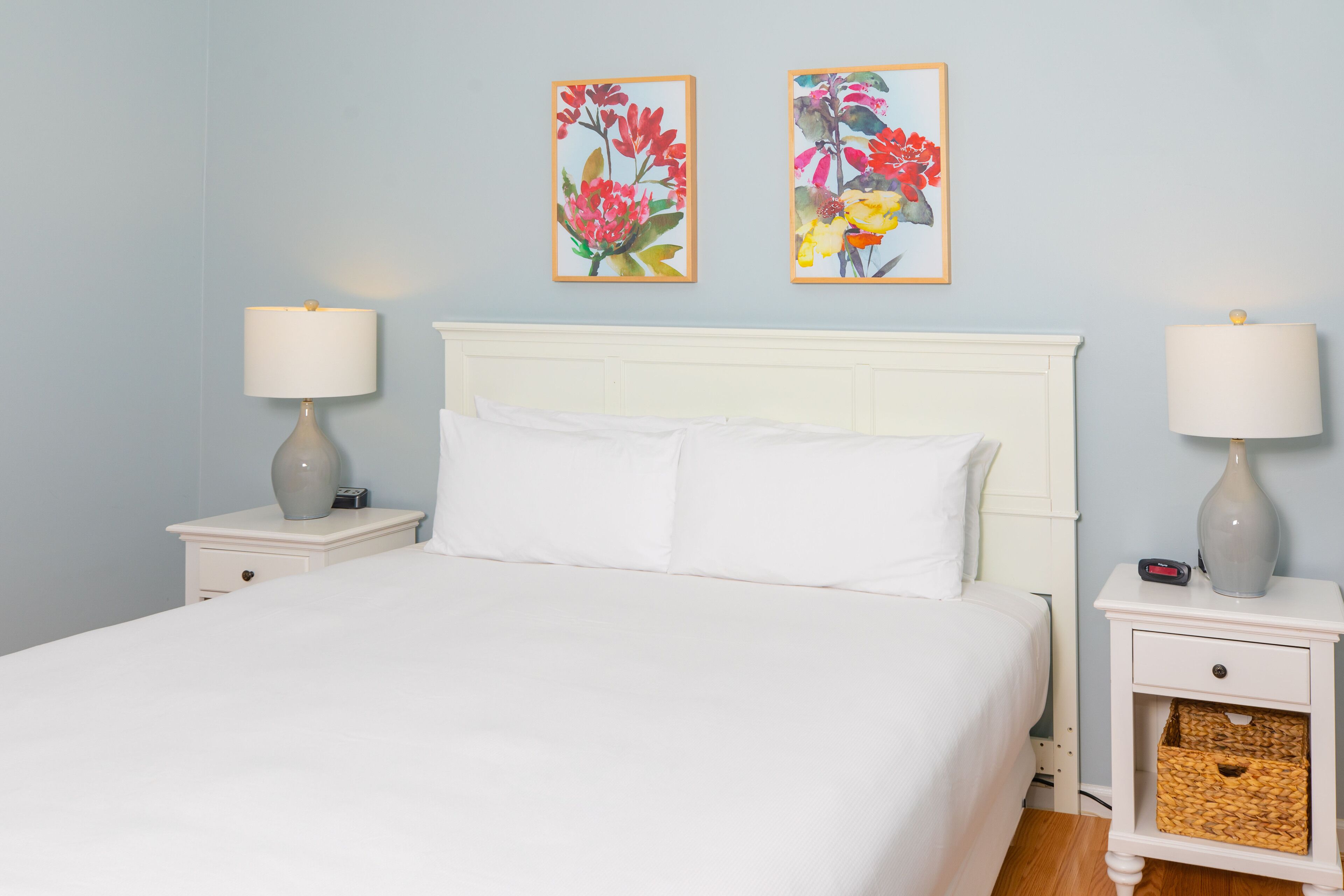 A neatly made bed with white linens, two bedside tables with lamps, and two framed floral paintings on the wall.