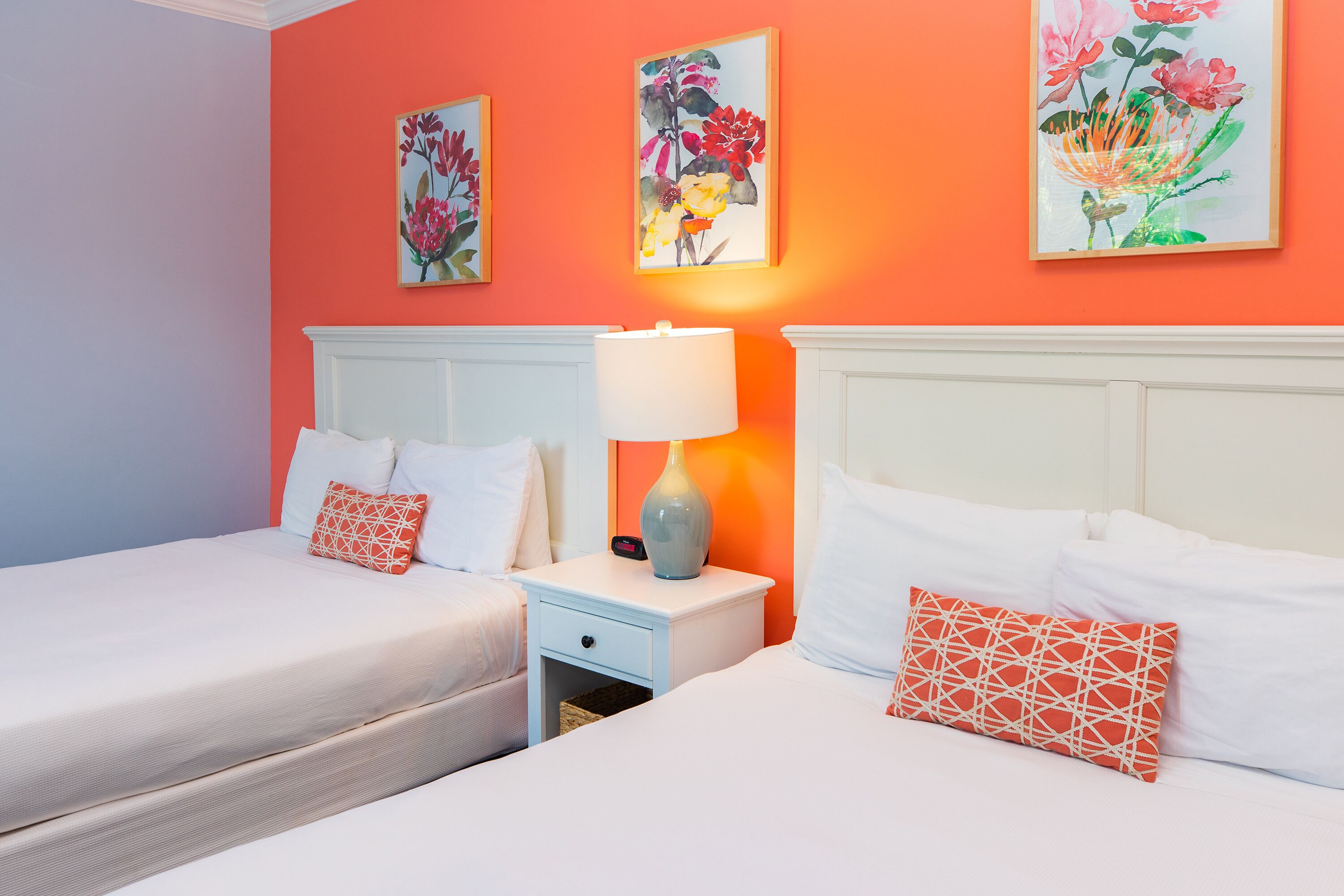 A hotel room with two beds, a nightstand with a lamp, and two floral paintings on the wall.